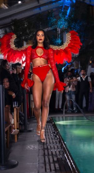 Model in red outfit with dramatic feathered wings performs on catwalk beside pool.