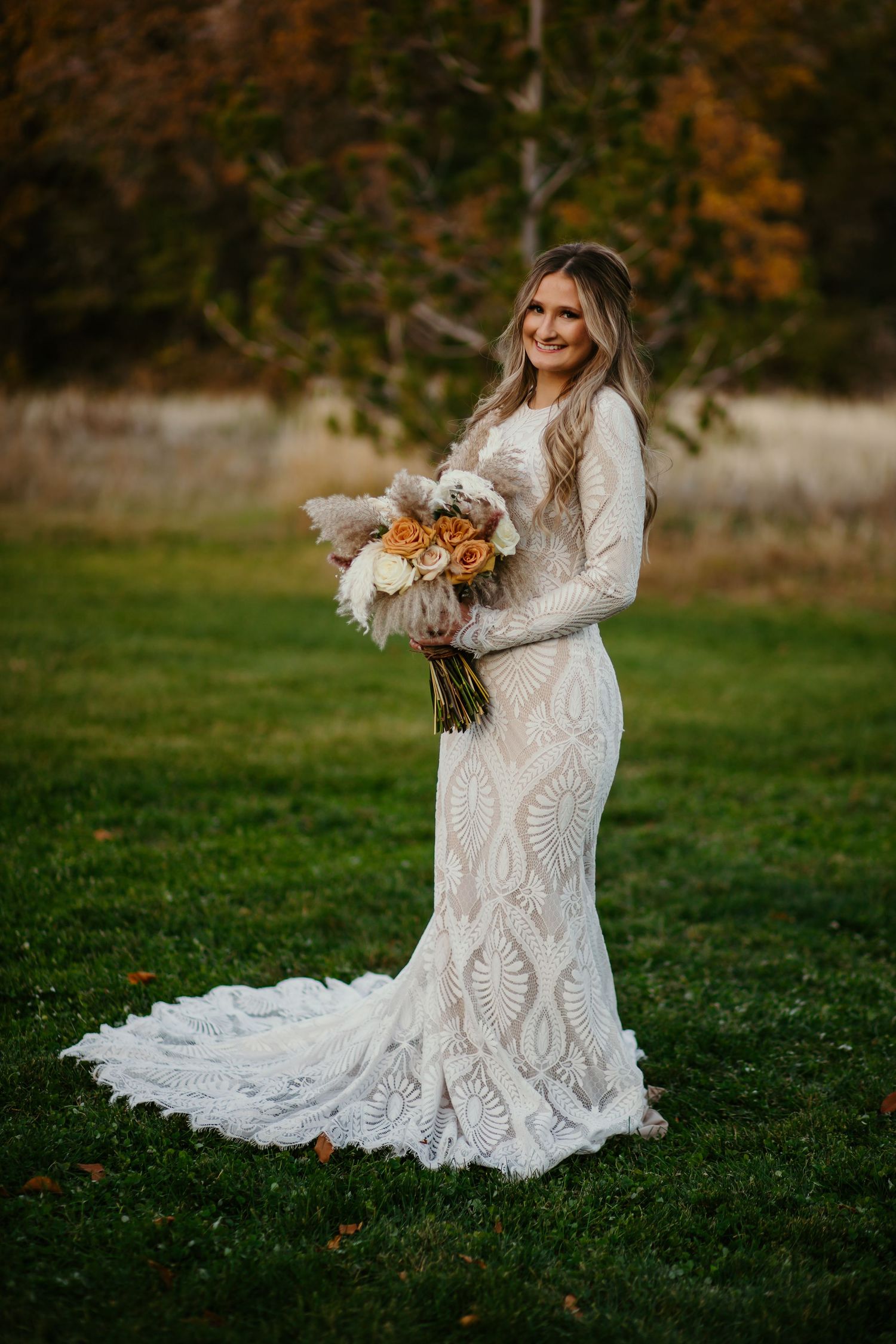 A bride in a lace wedding dress poses with her bouquet in an autumn field.