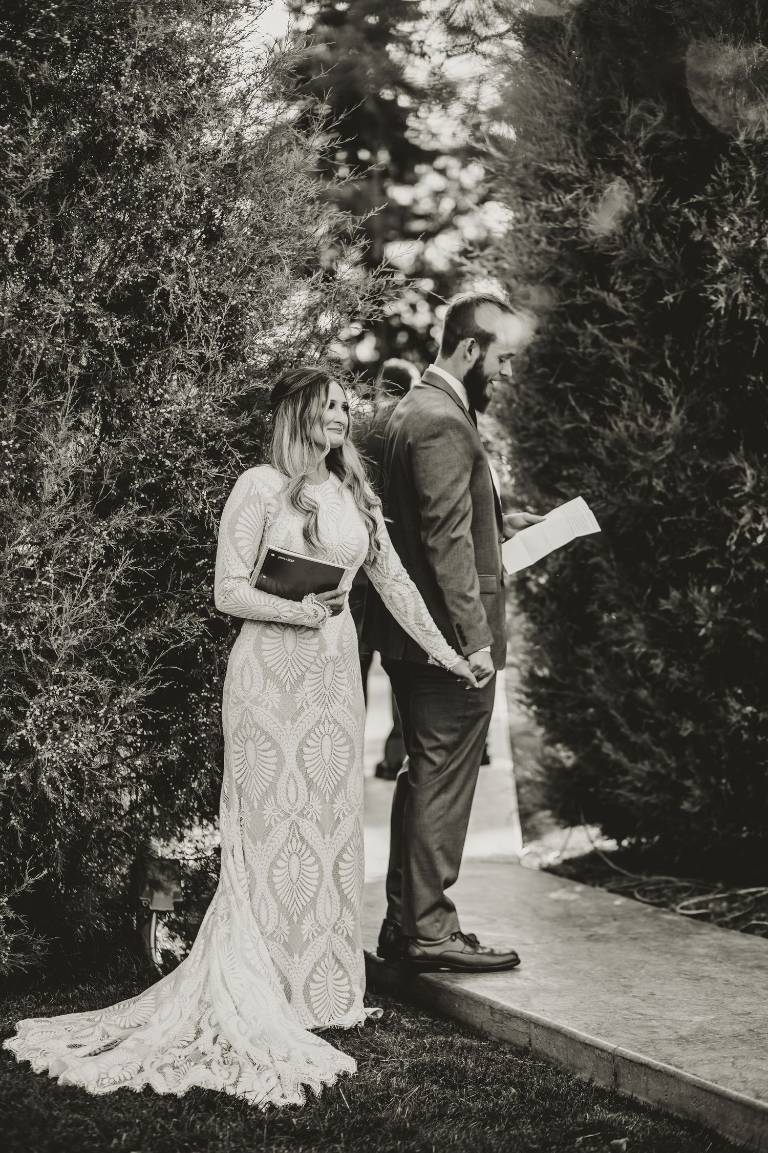 Black and white photo of couple in wedding attire standing on garden path during ceremony.