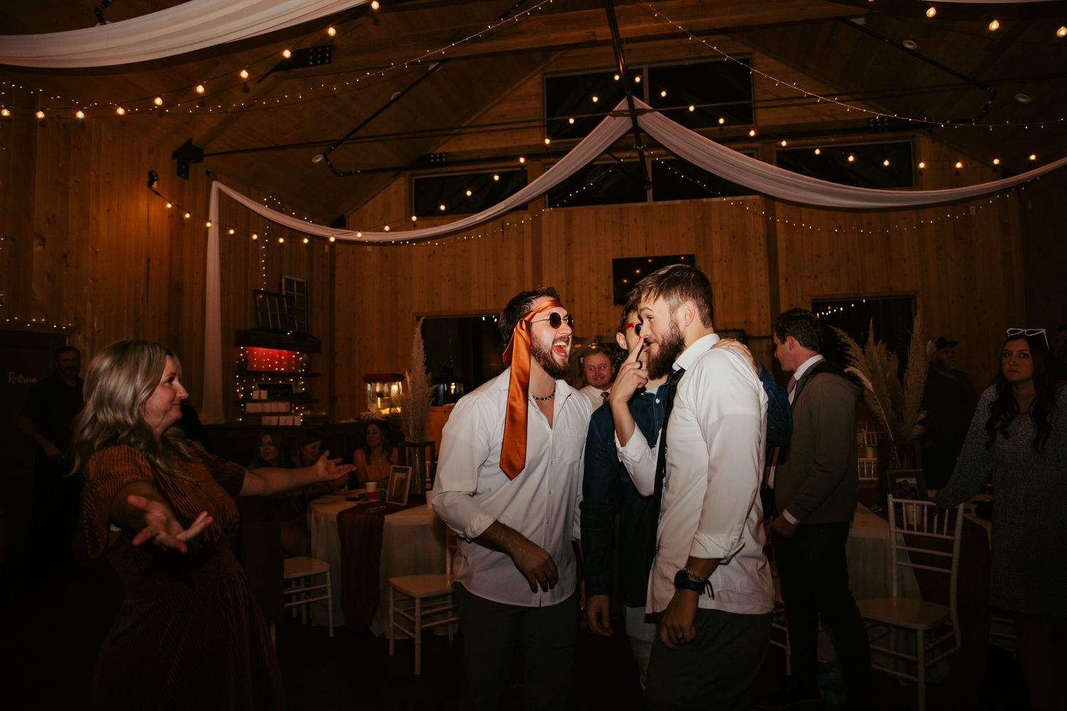 Wedding reception guests mingling under string lights in rustic barn venue.