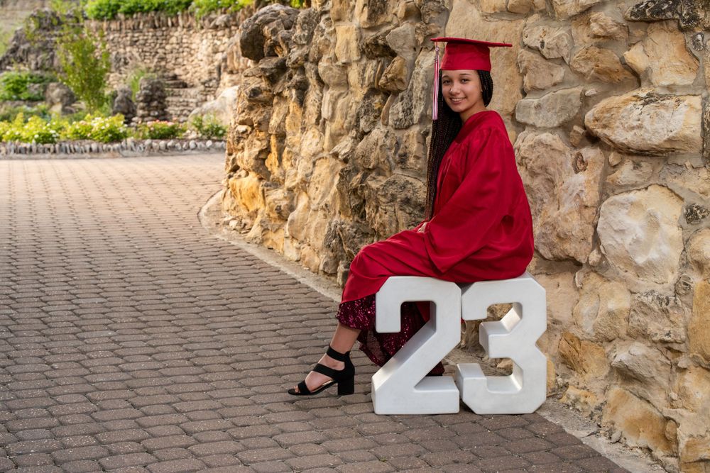 Graduate in red cap and gown sits on decorative numbers 23 against stone wall.