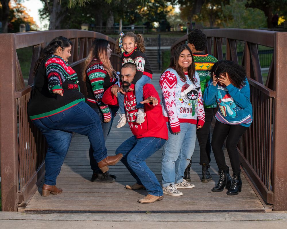 Group wearing festive Christmas sweaters poses playfully on a wooden bridge.