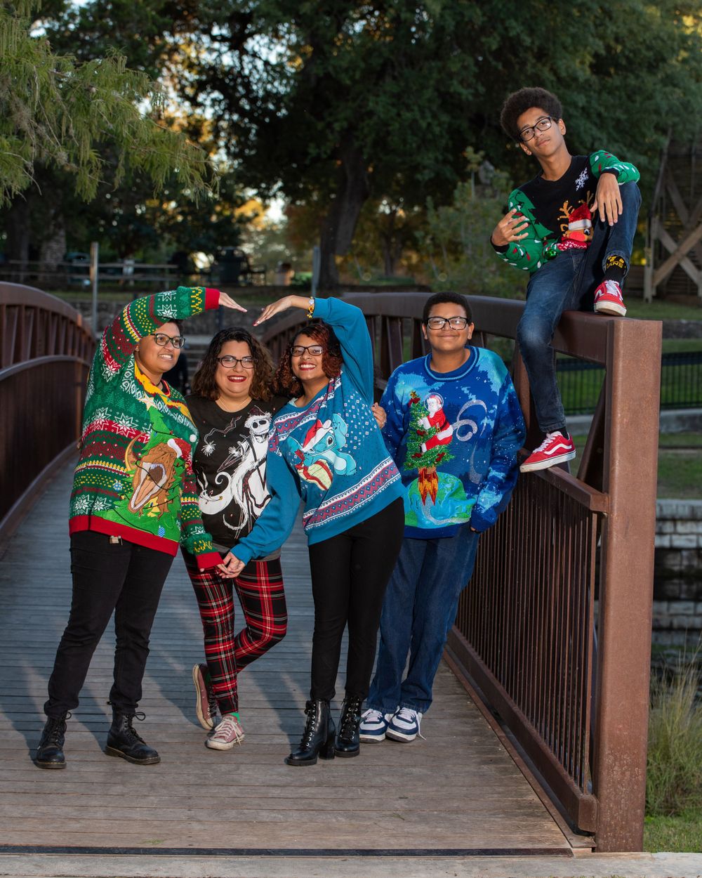 Friends in holiday sweaters strike fun poses on a wooden bridge at sunset.