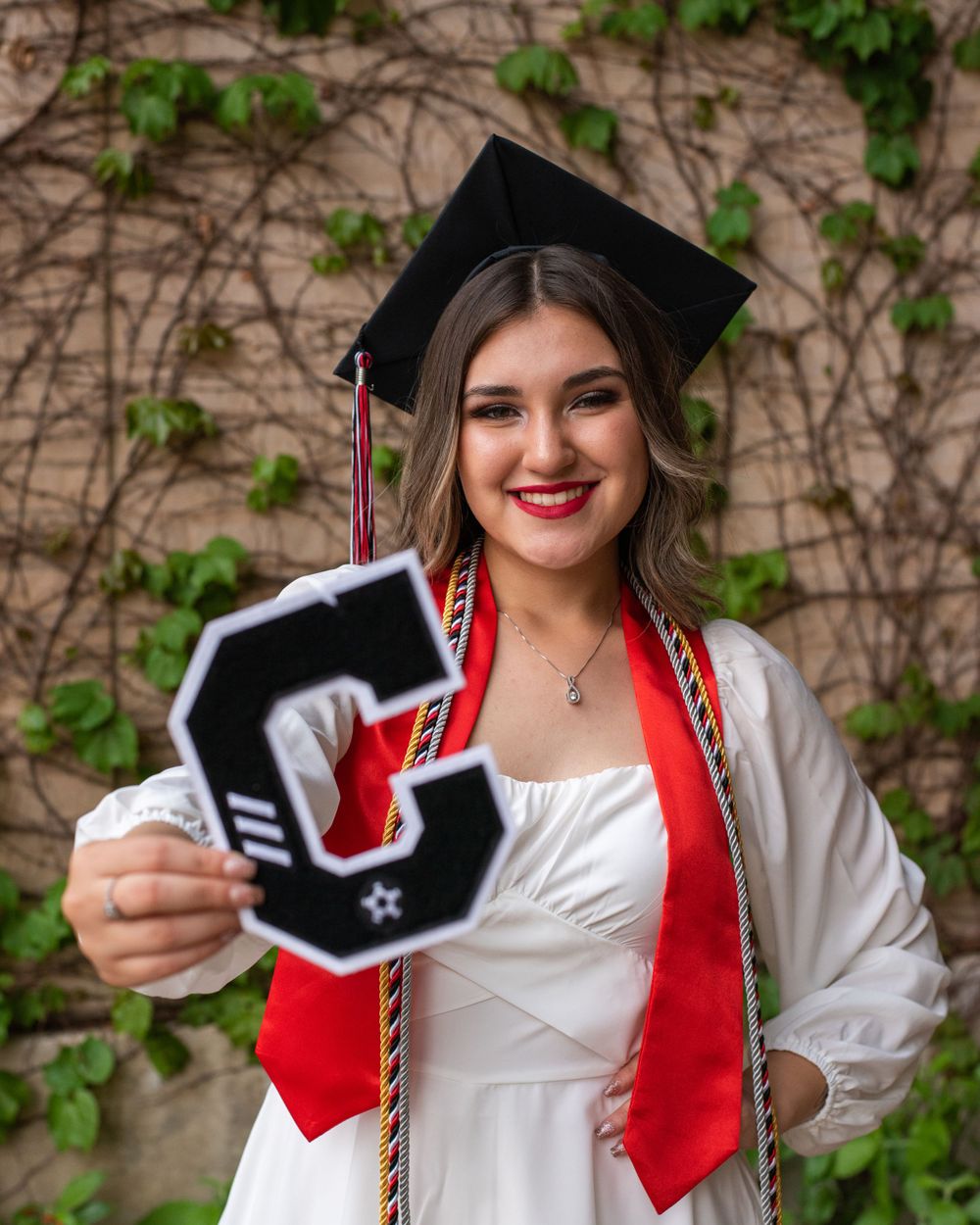Graduate in cap and gown holds up decorative letter G against ivy wall.