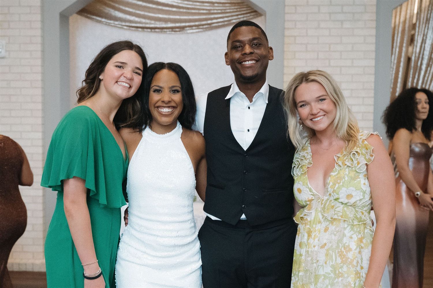 A group of four friends dressed in formal attire smile together at a special event with light colored walls behind them.