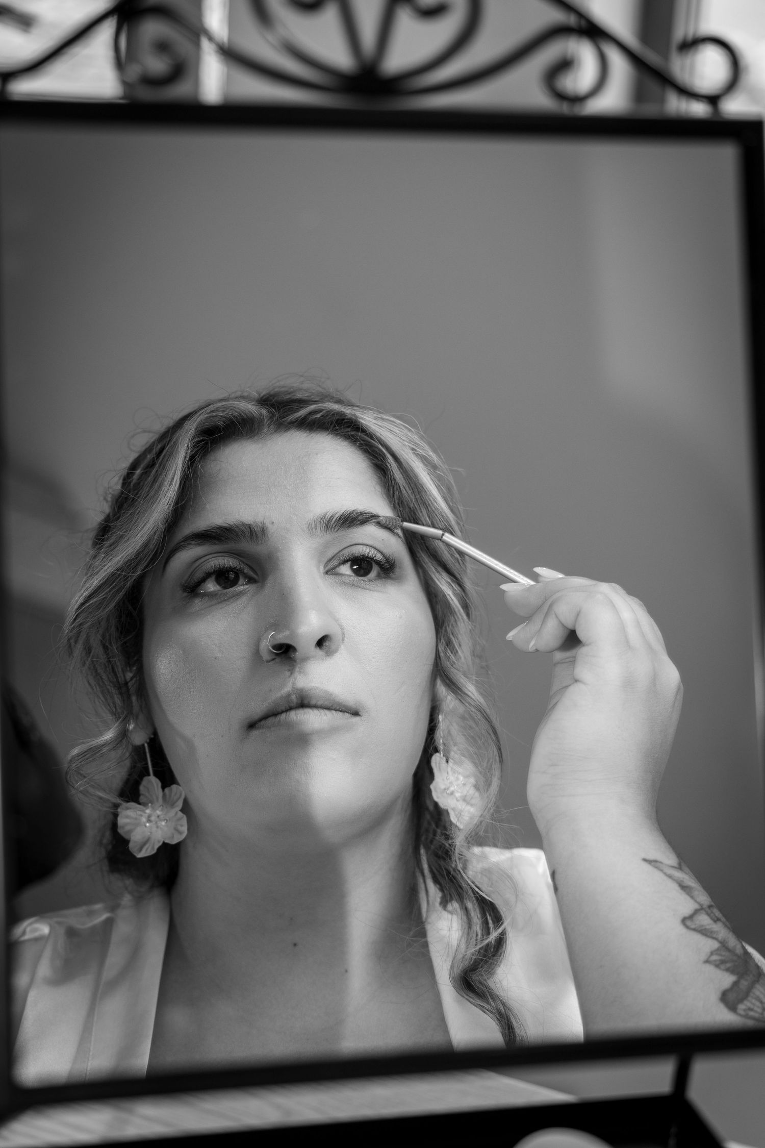 Black and white portrait of a bride grooming her eyebrows in a decorative mirror.