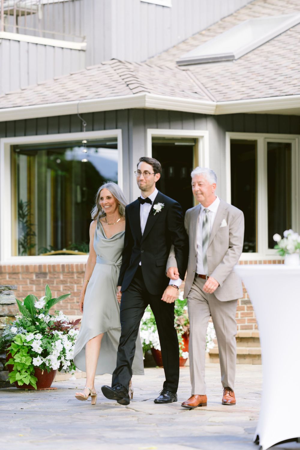 Three people in formal attire walk together outside a white house with brick accents.