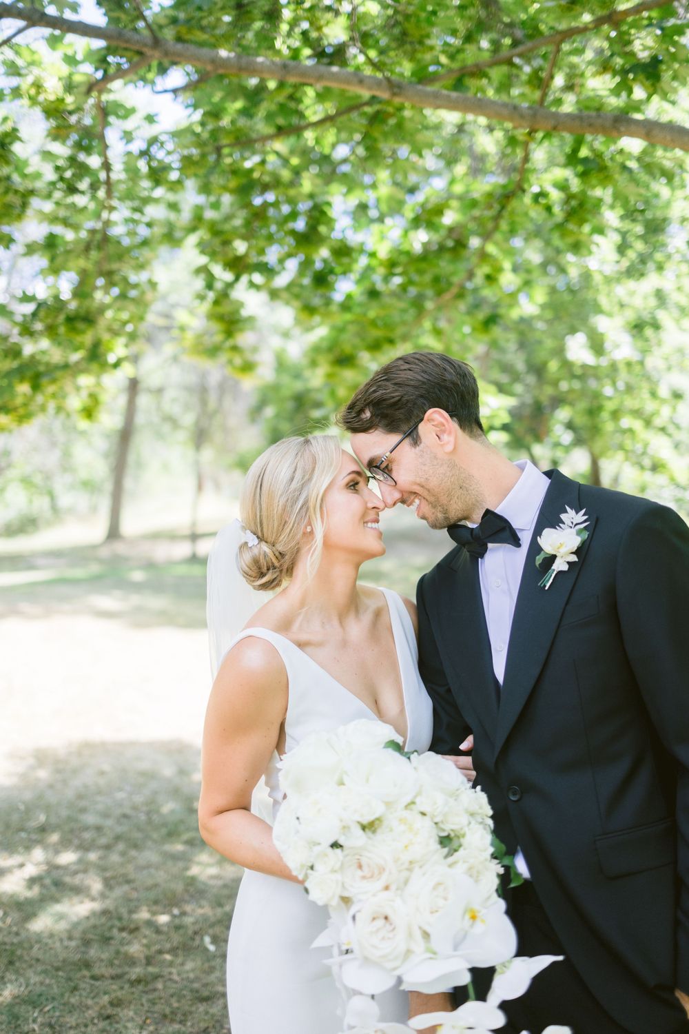 Romantic moment captured under green trees with white floral bouquet.