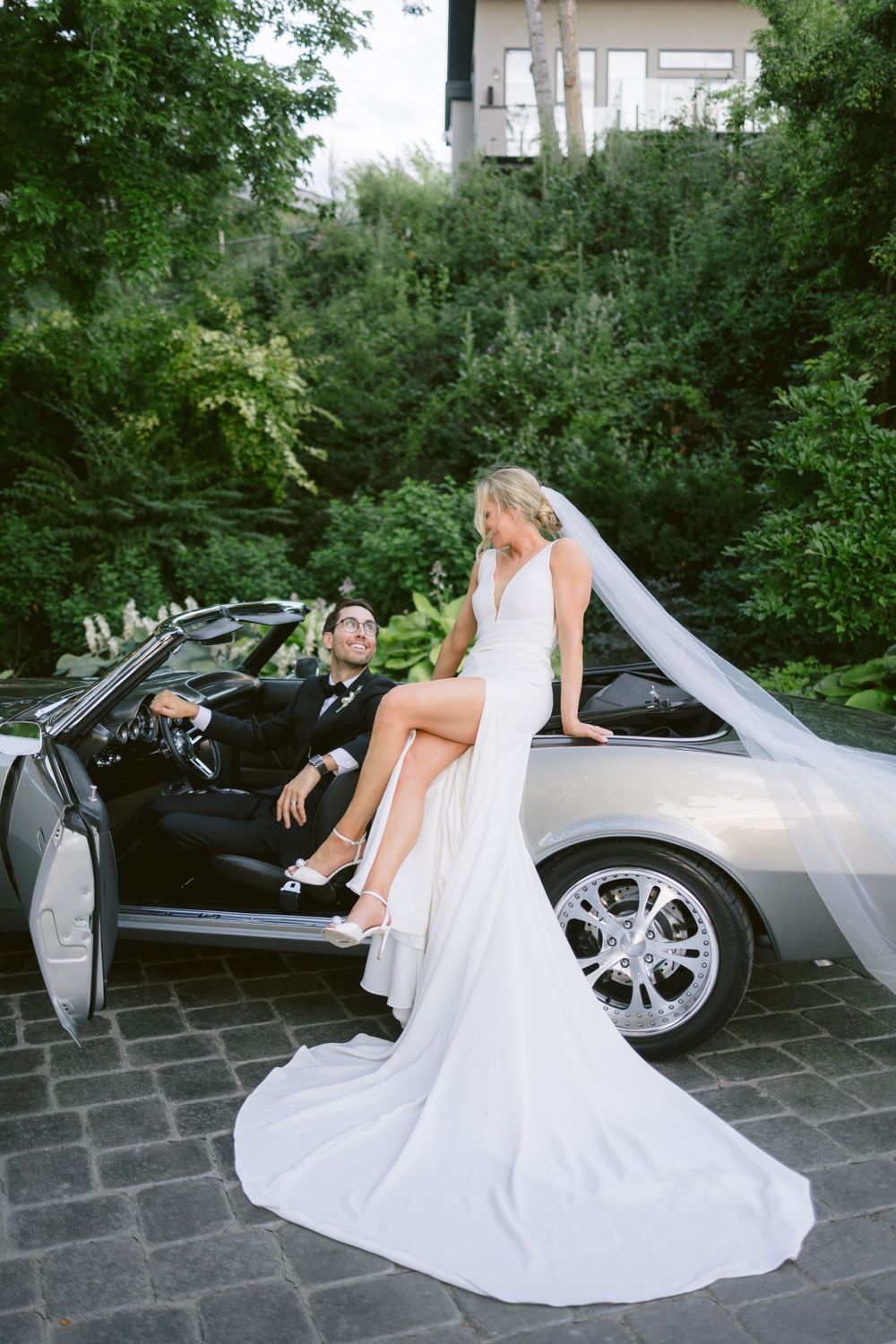 Bride in flowing white gown poses on silver convertible while groom sits in driver's seat.