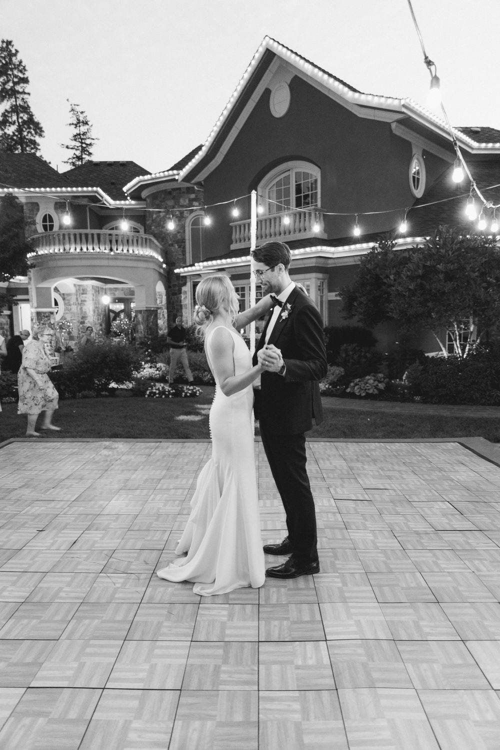 A romantic black and white wedding photo showing a couple dancing together on a patio in front of an elegant lit mansion.