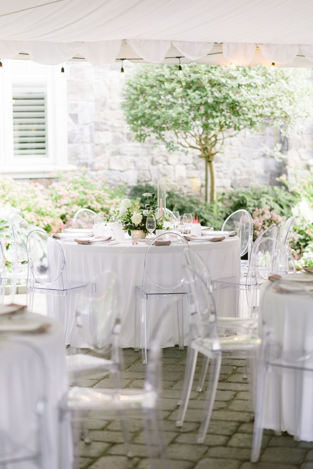 Elegant outdoor wedding reception setup with white tablecloths, clear ghost chairs and garden views in the background.