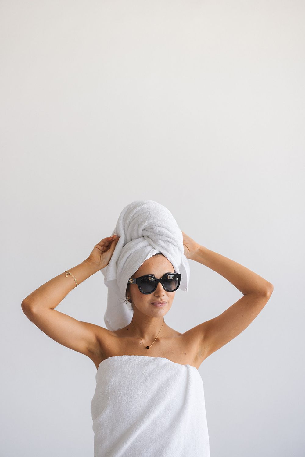 Person wrapped in white towels making playful poses against a light background while wearing sunglasses.