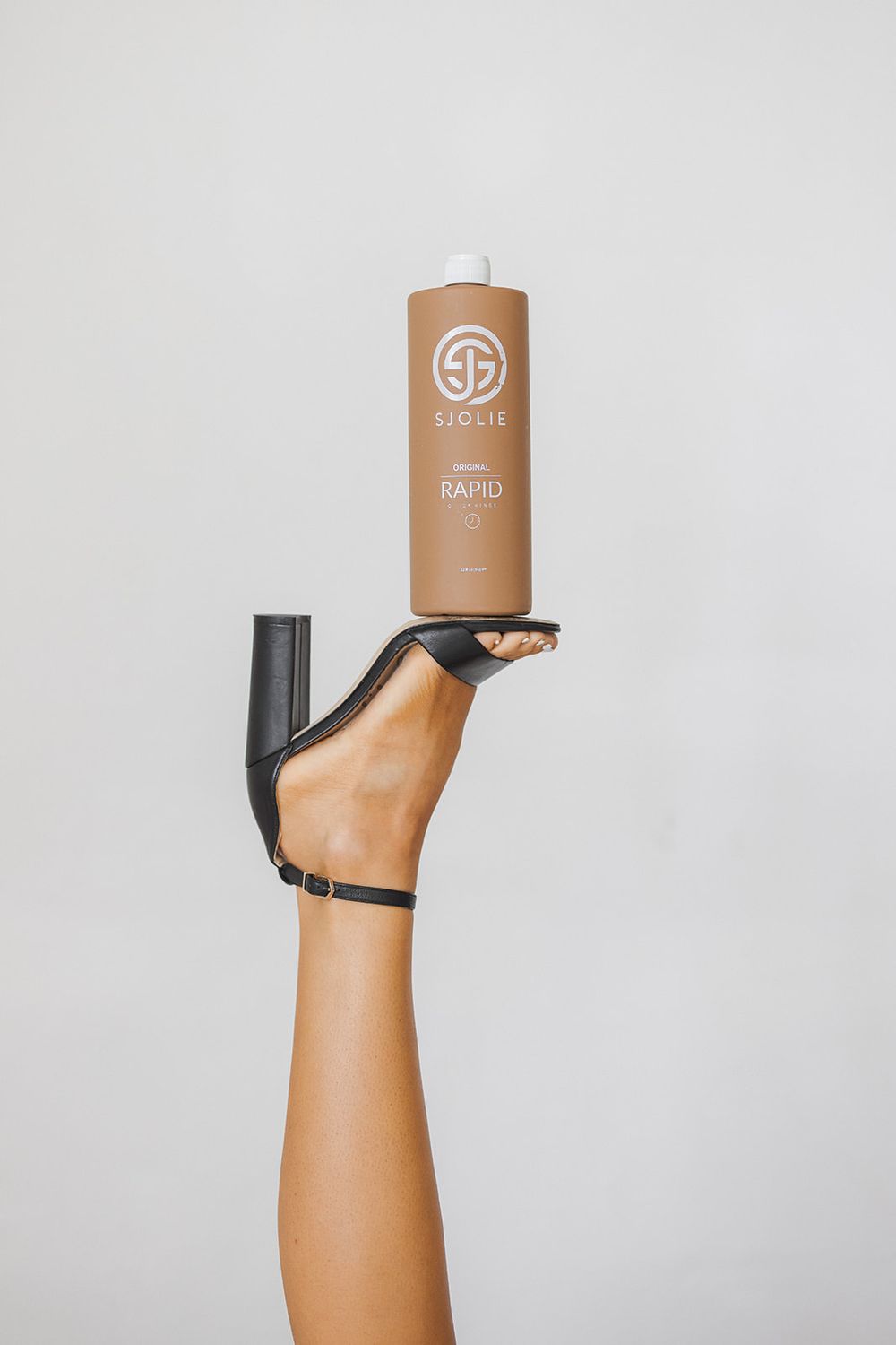 A tan skinned foot wearing a black high heel shoe balances a brown cylindrical bottle on its sole against a gray background.
