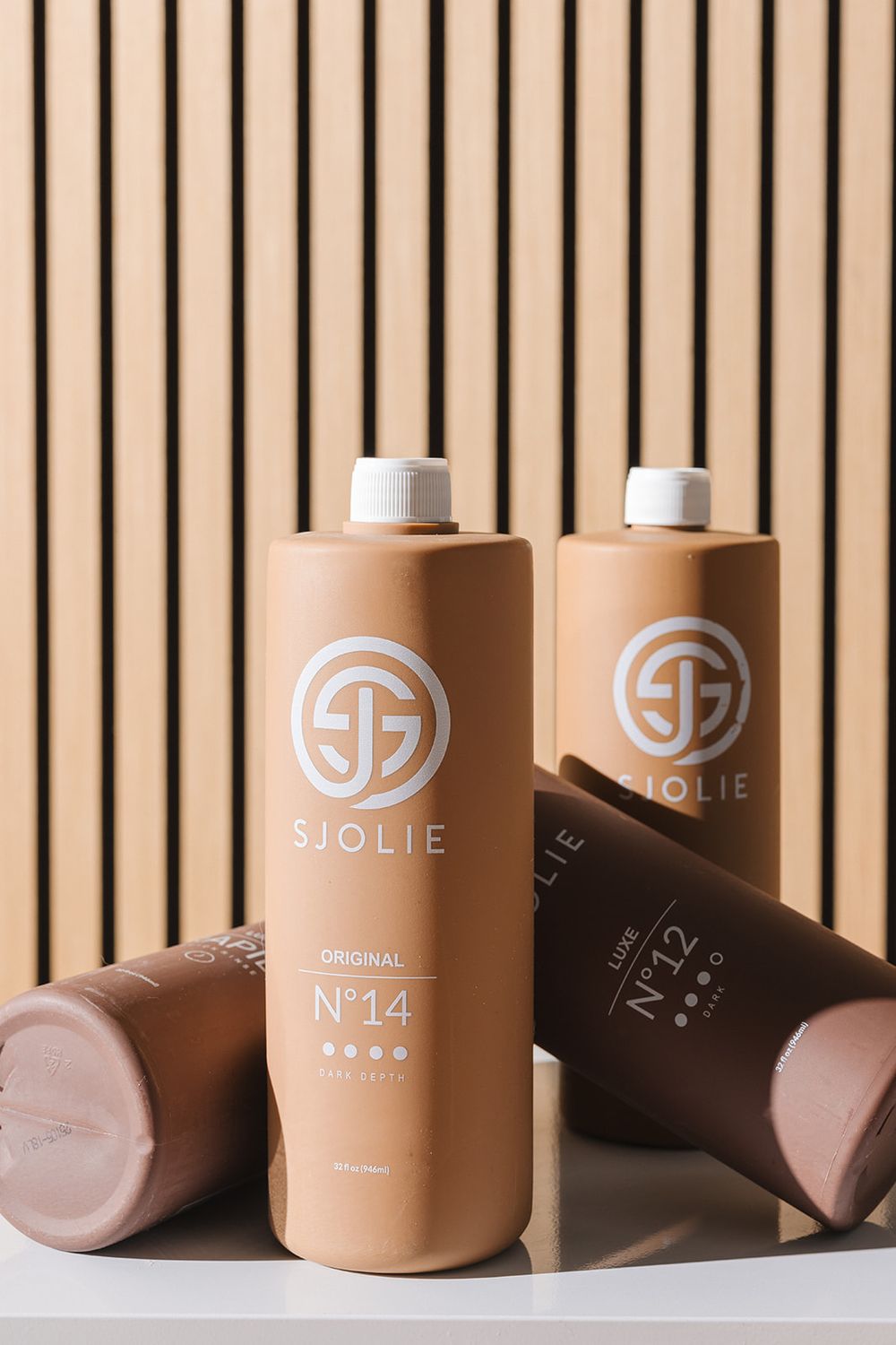 Three Sjolie tanning lotion bottles with a beige colored body and white caps displayed against a wooden slat background.