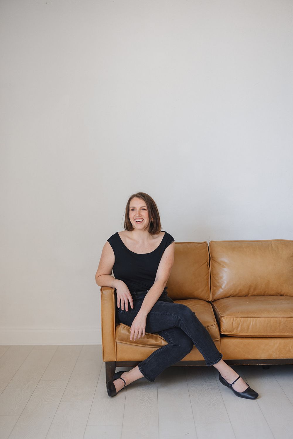 A person in a black top and jeans sits casually on a tan leather couch against a light gray wall.