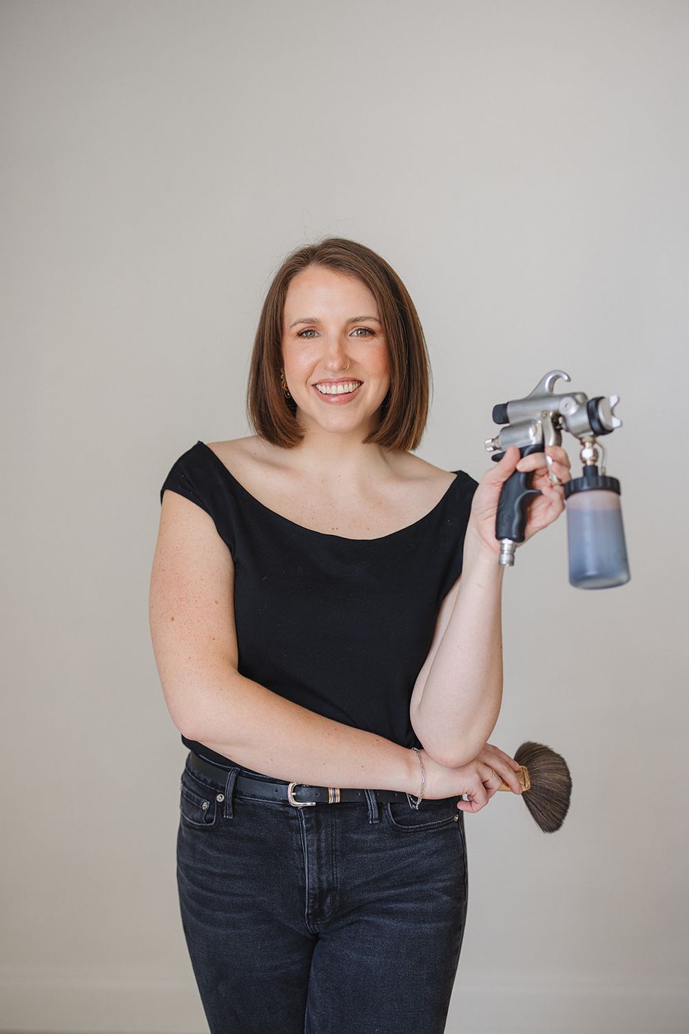 Person in black top and jeans holding a spray paint gun against light gray background.