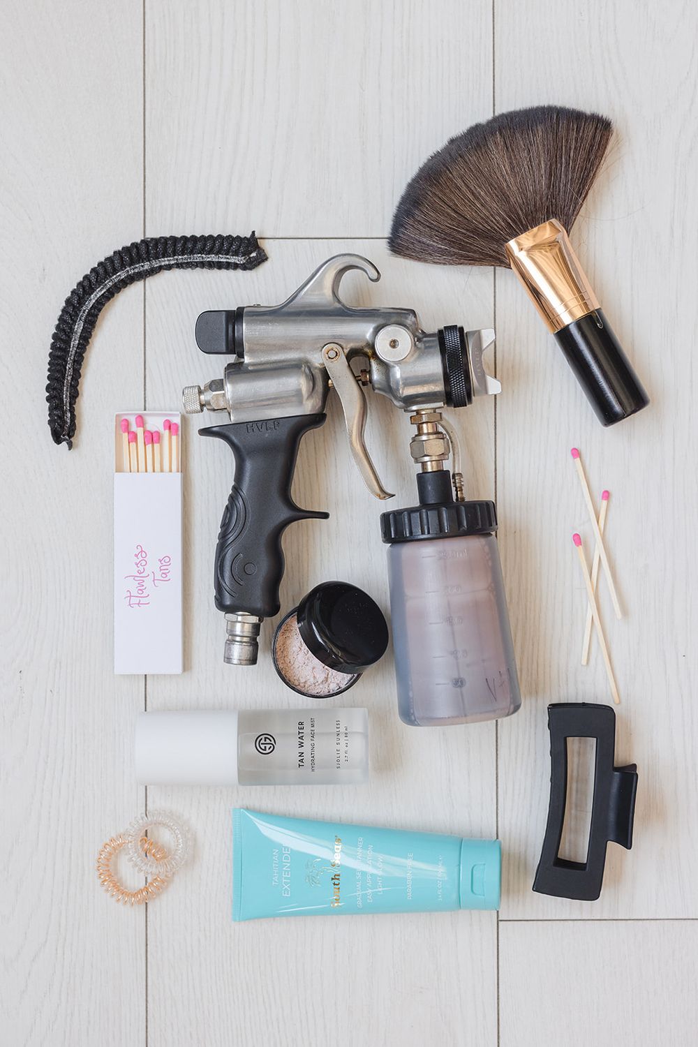 Professional spray tan equipment including gun, solution bottles, cosmetic brush and skincare products laid out on light wooden surface.