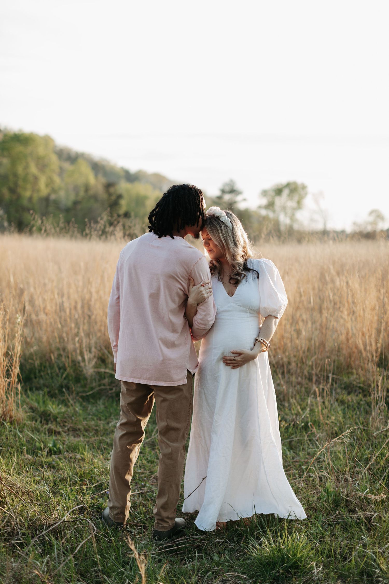 An intimate maternity photo of a couple in a sunlit field of tall grass.