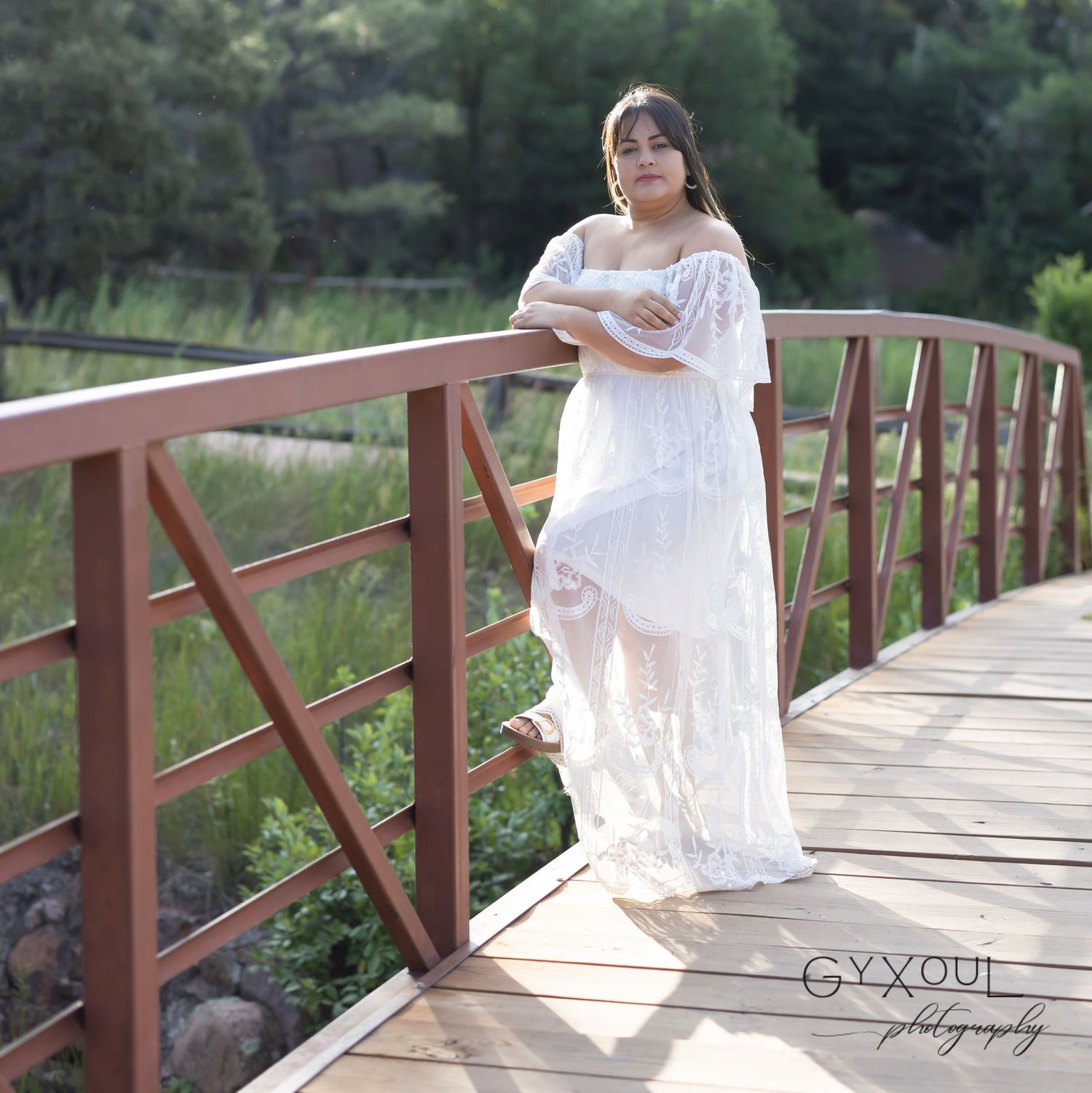 Person in flowing white dress poses on rustic wooden bridge in nature setting.