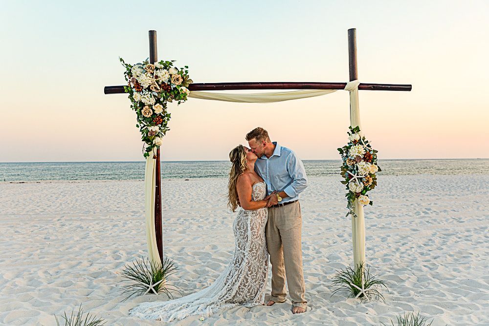 A romantic beach wedding ceremony setup with a wooden arch decorated with white flowers during sunset.