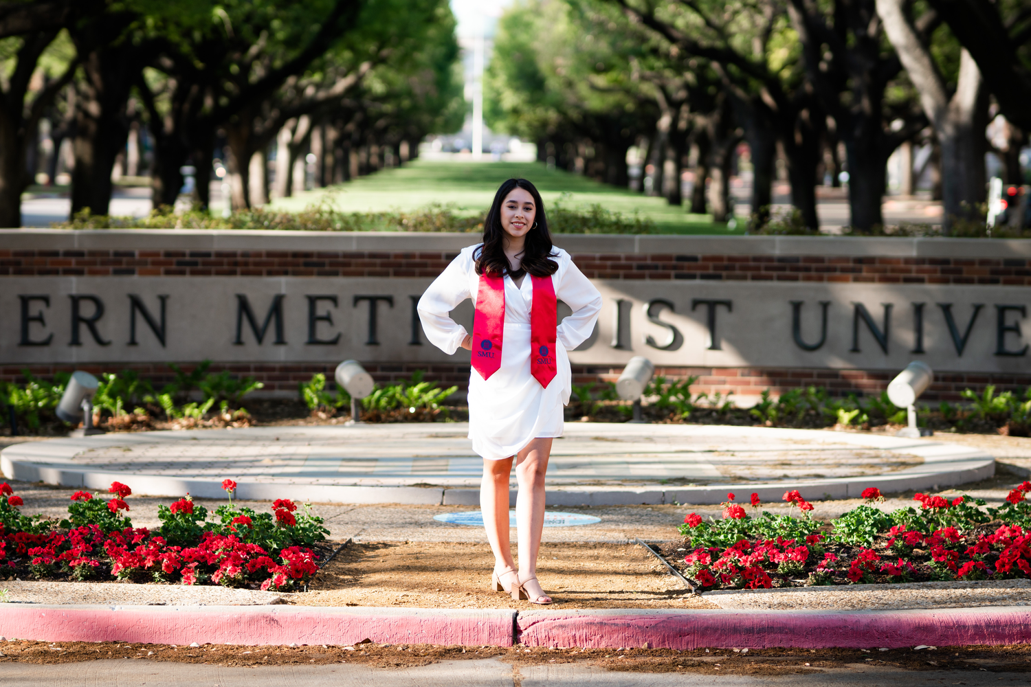 Graduate in red and white regalia poses at Southern Methodist University entrance.