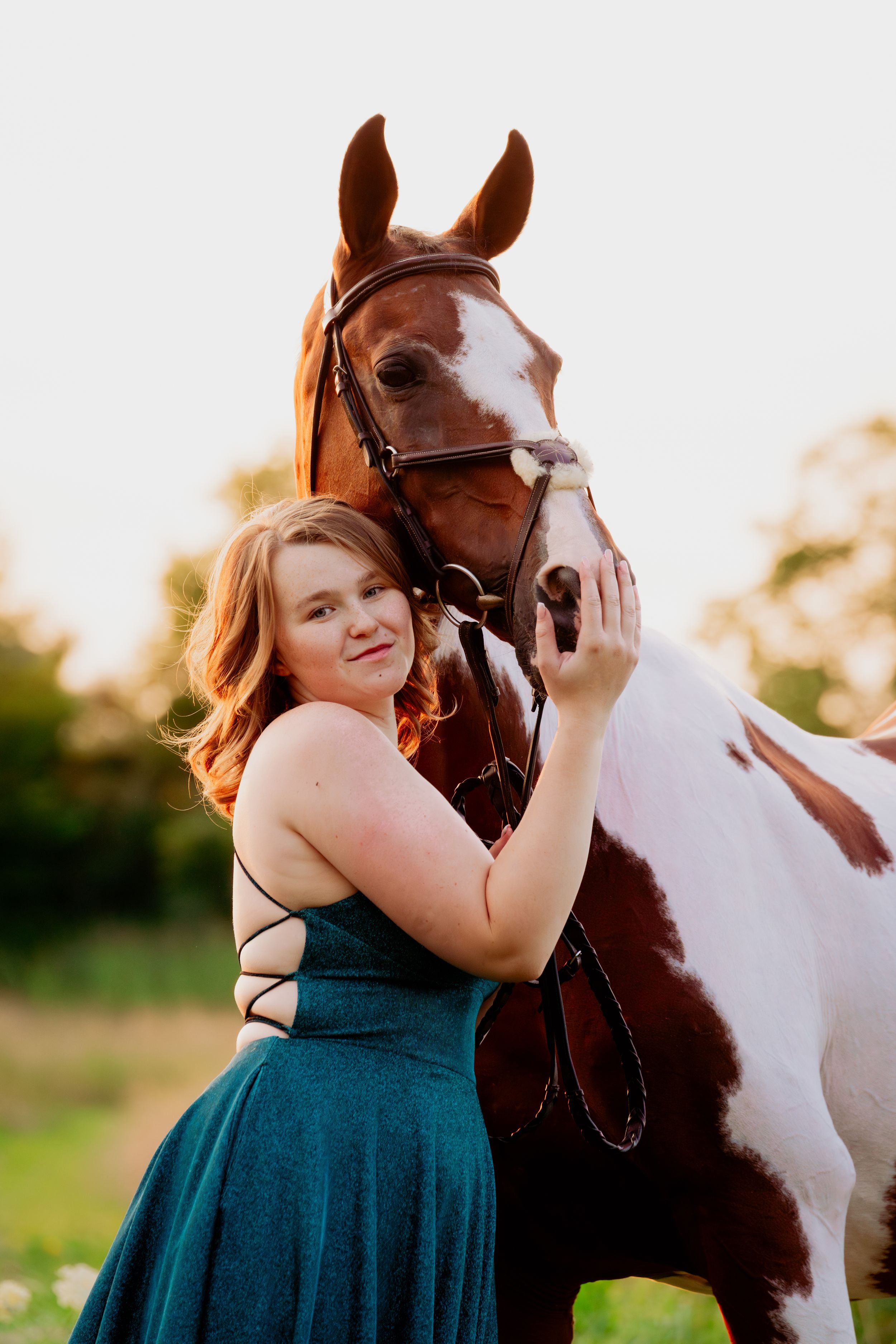 A person in a teal dress embraces a brown and white horse during an outdoor photoshoot at sunset.