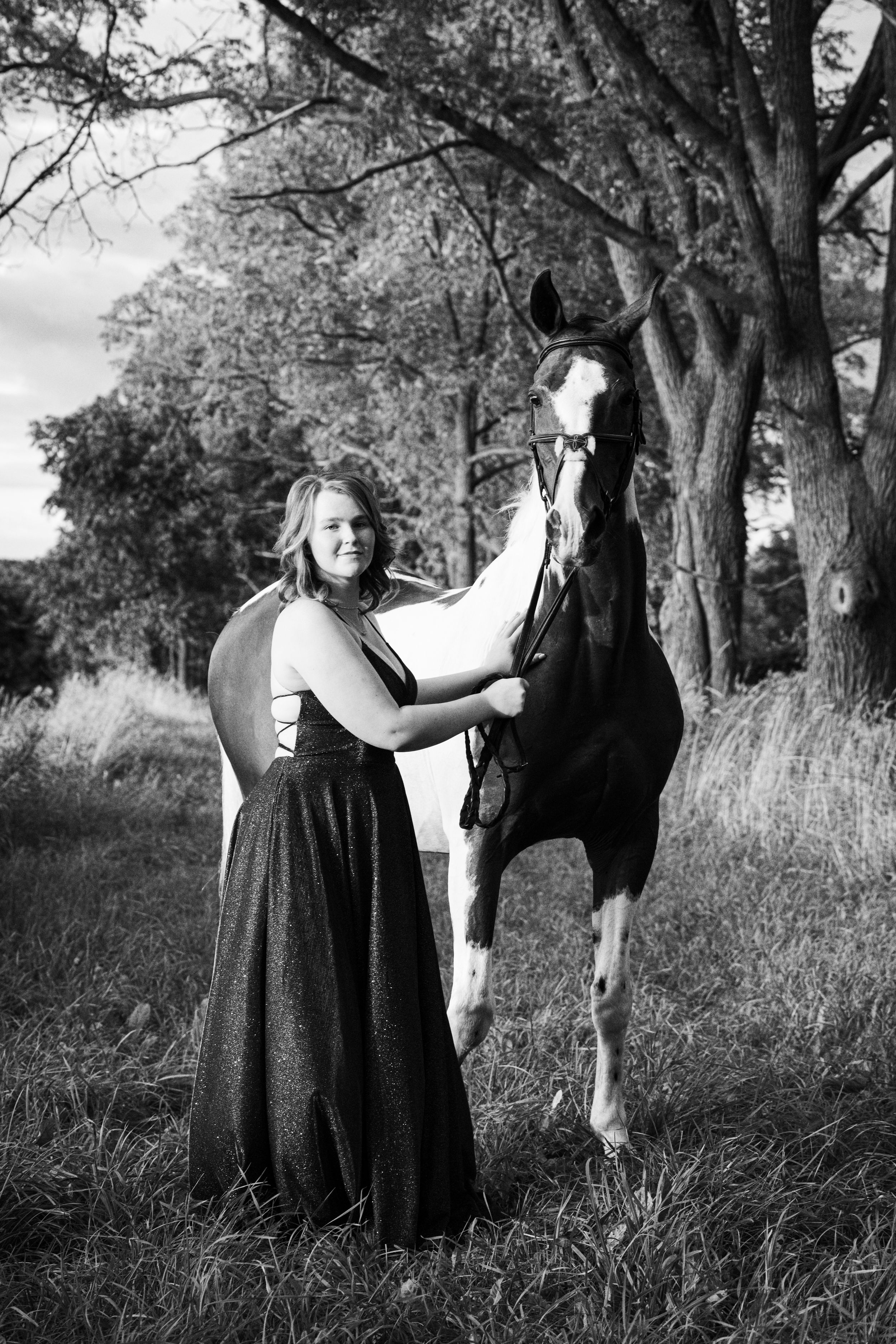 A black and white portrait of a person in a dress standing with a paint horse in a wooded outdoor setting.