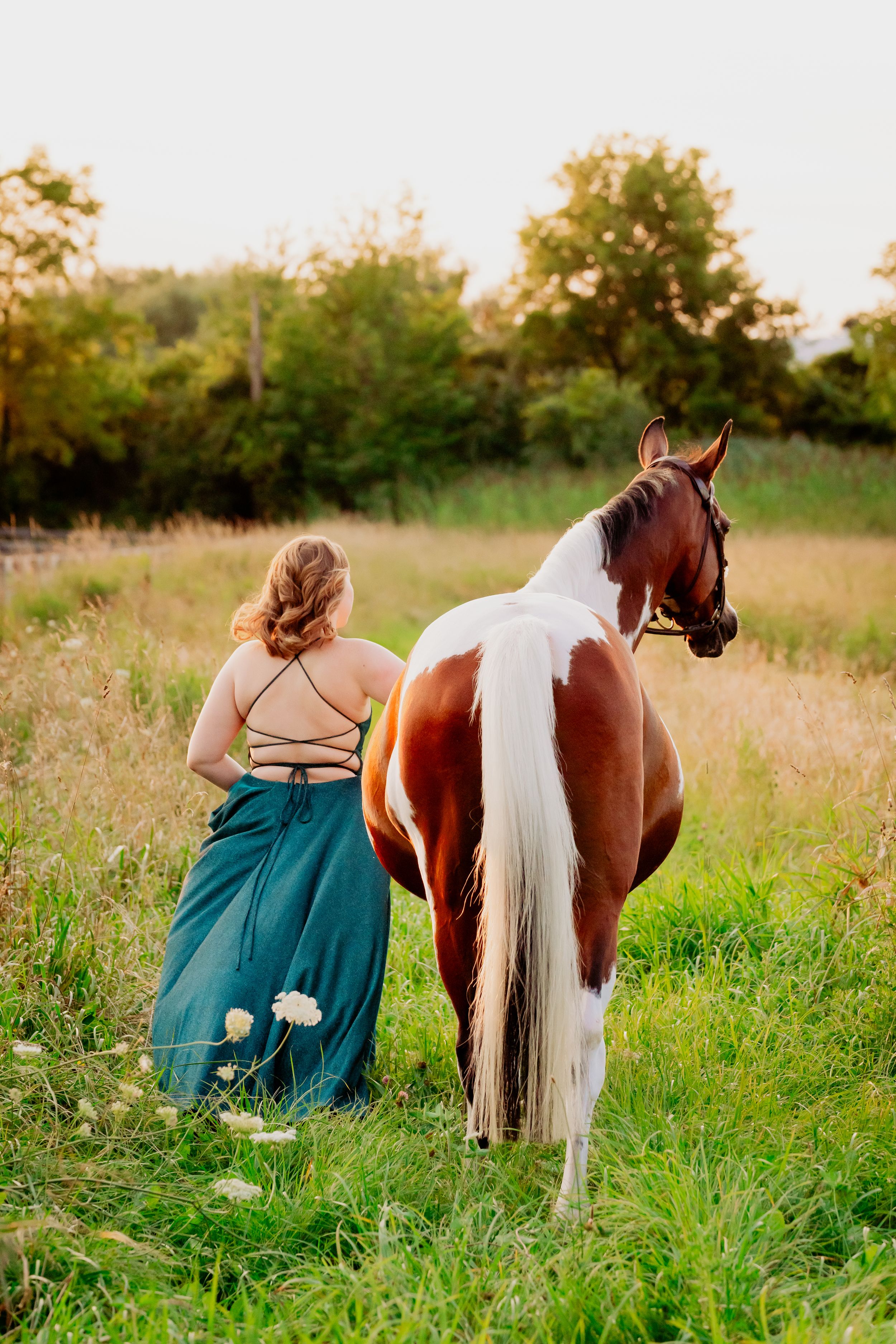 A person in a flowing dress walks with their paint horse through a golden summer field.
