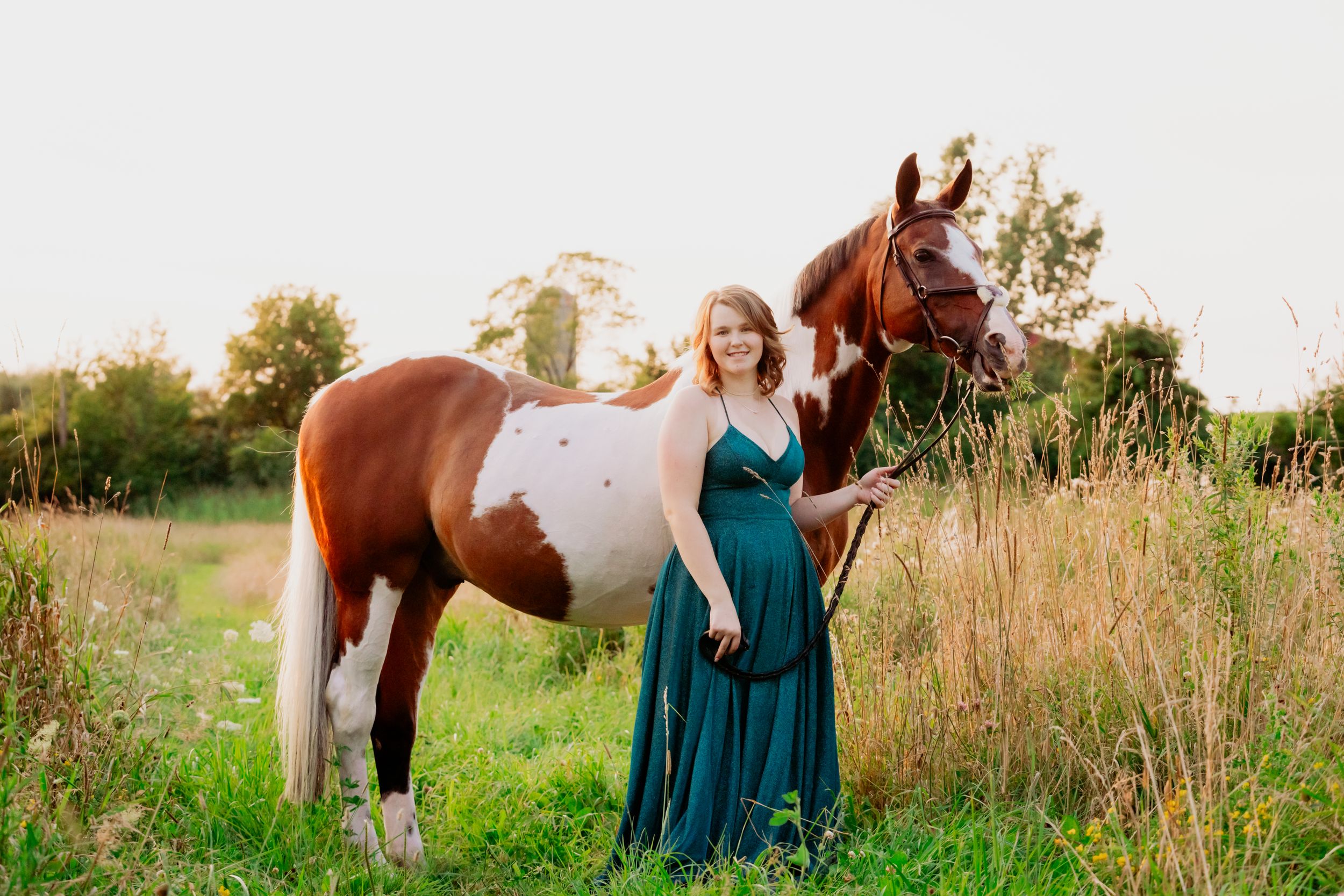 A person in a teal dress stands beside a paint horse in a sunlit meadow during summer.