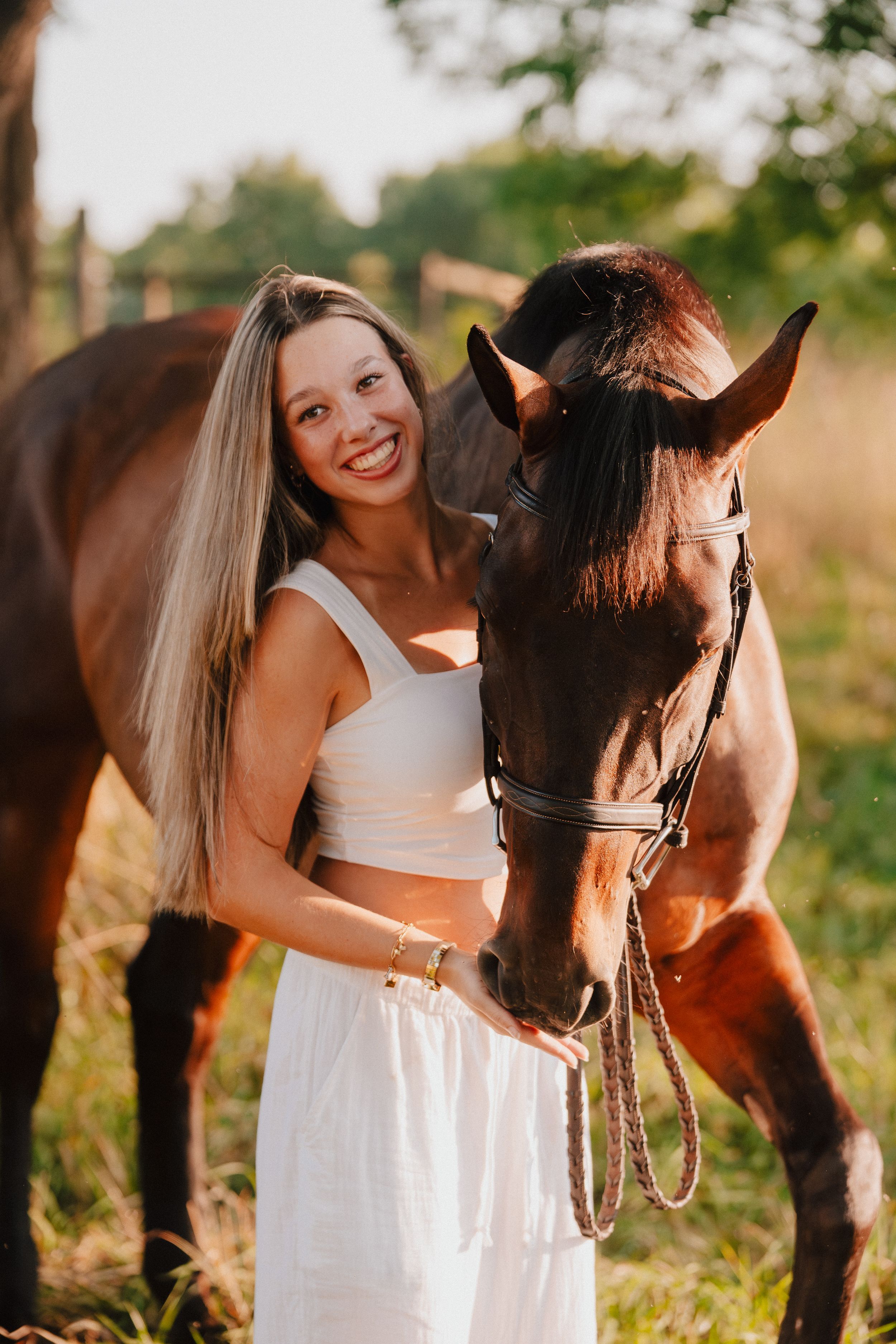 A beautiful bay horse and person in a white dress stand together in a sun-dappled field at golden hour.