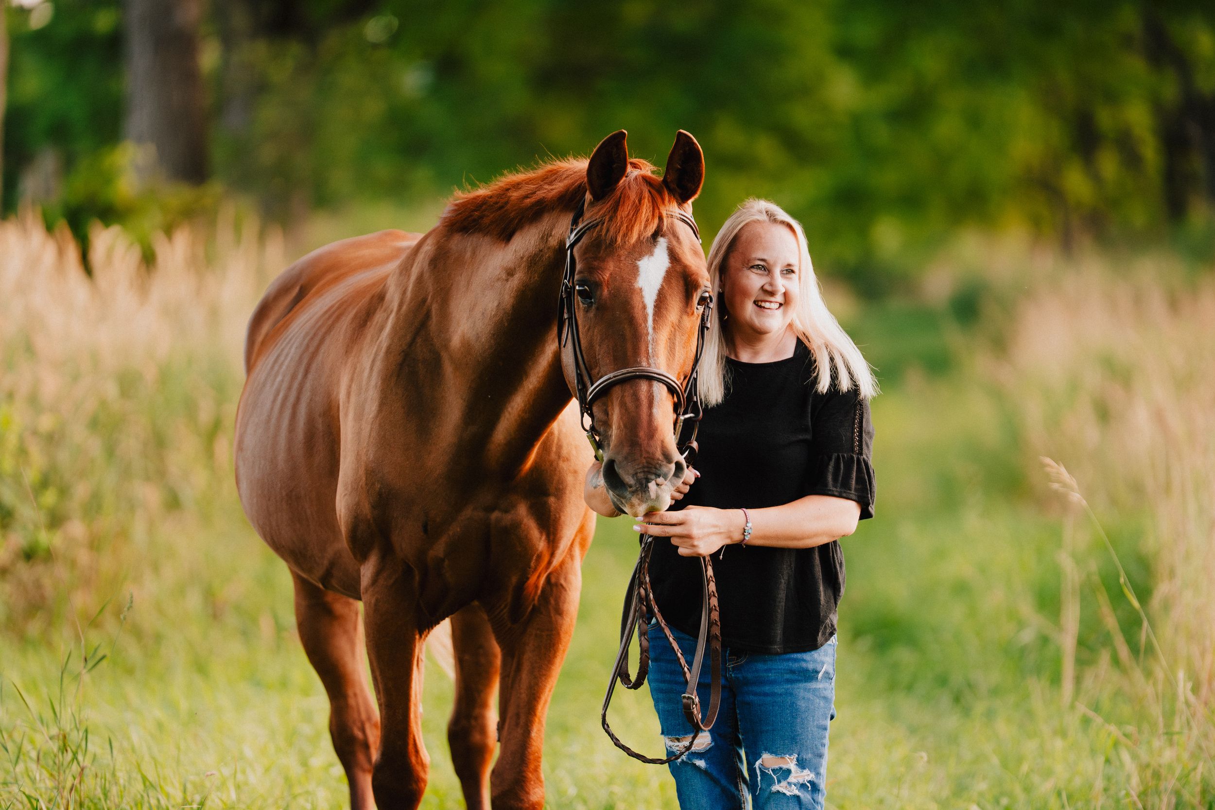 Person in jeans and black shirt leads brown horse through green summer meadow at sunset.