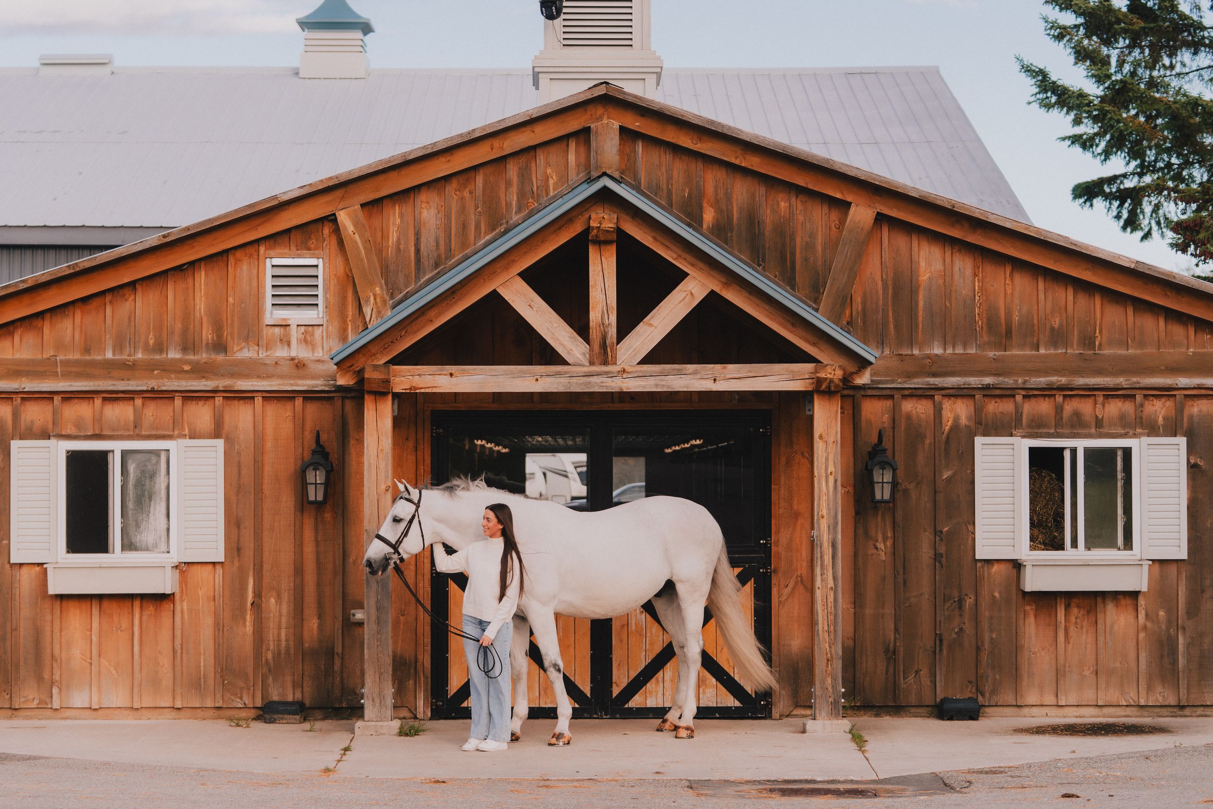 A white horse stands outside a rustic wooden barn with cedar siding and a triangular entrance overhang.