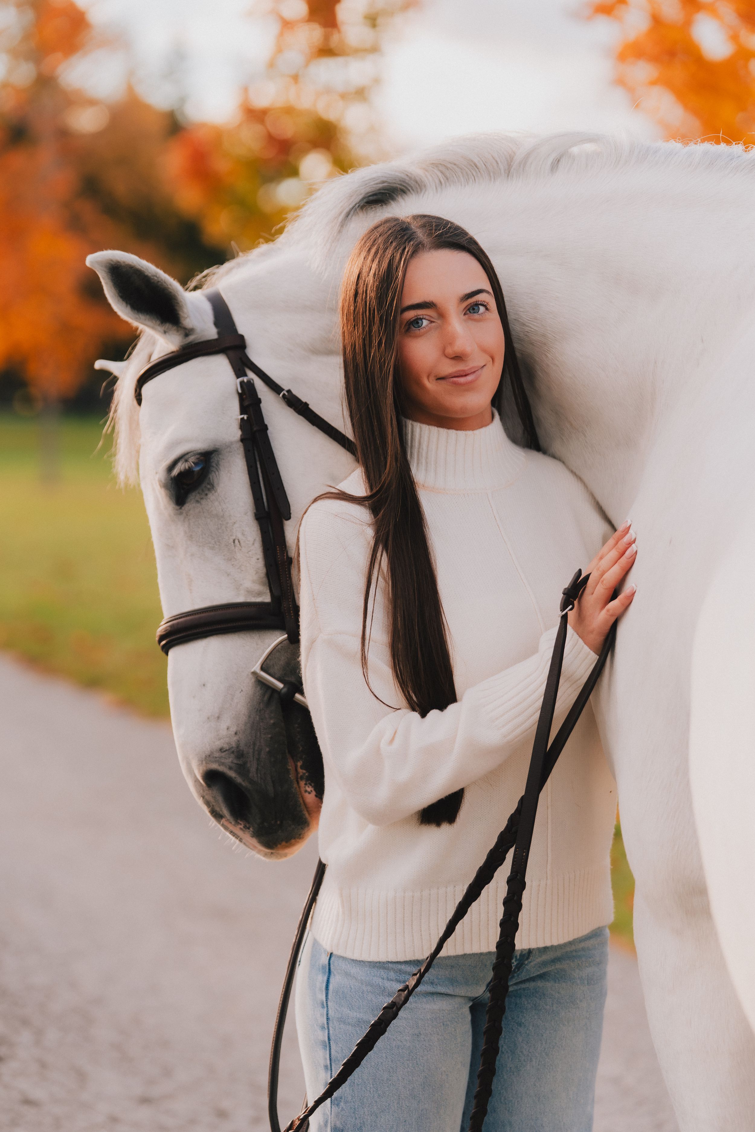 A person in a white sweater stands next to a beautiful white horse against vibrant fall foliage in the background.
