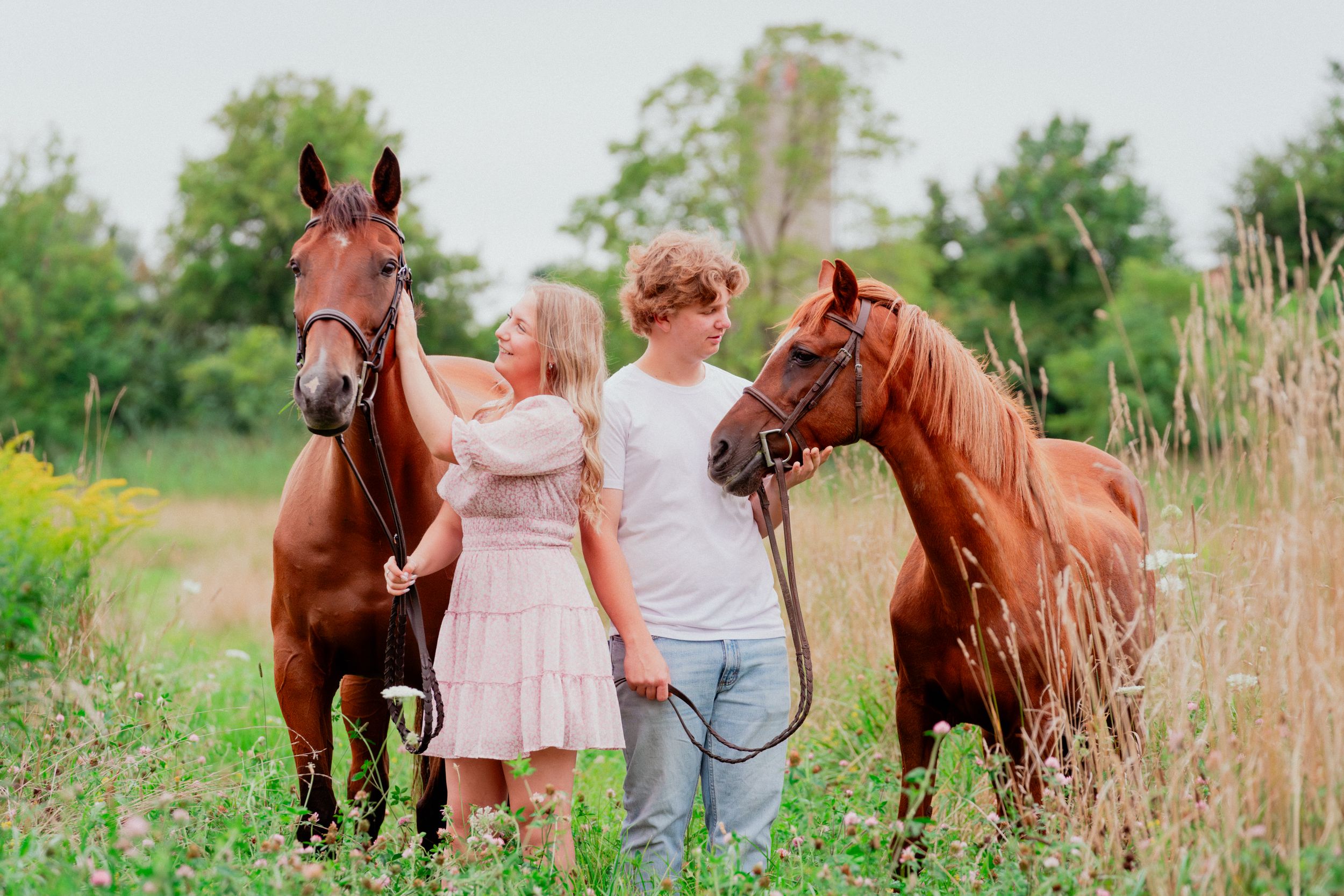 Two people pose with their chestnut horses in a sunny summer field.