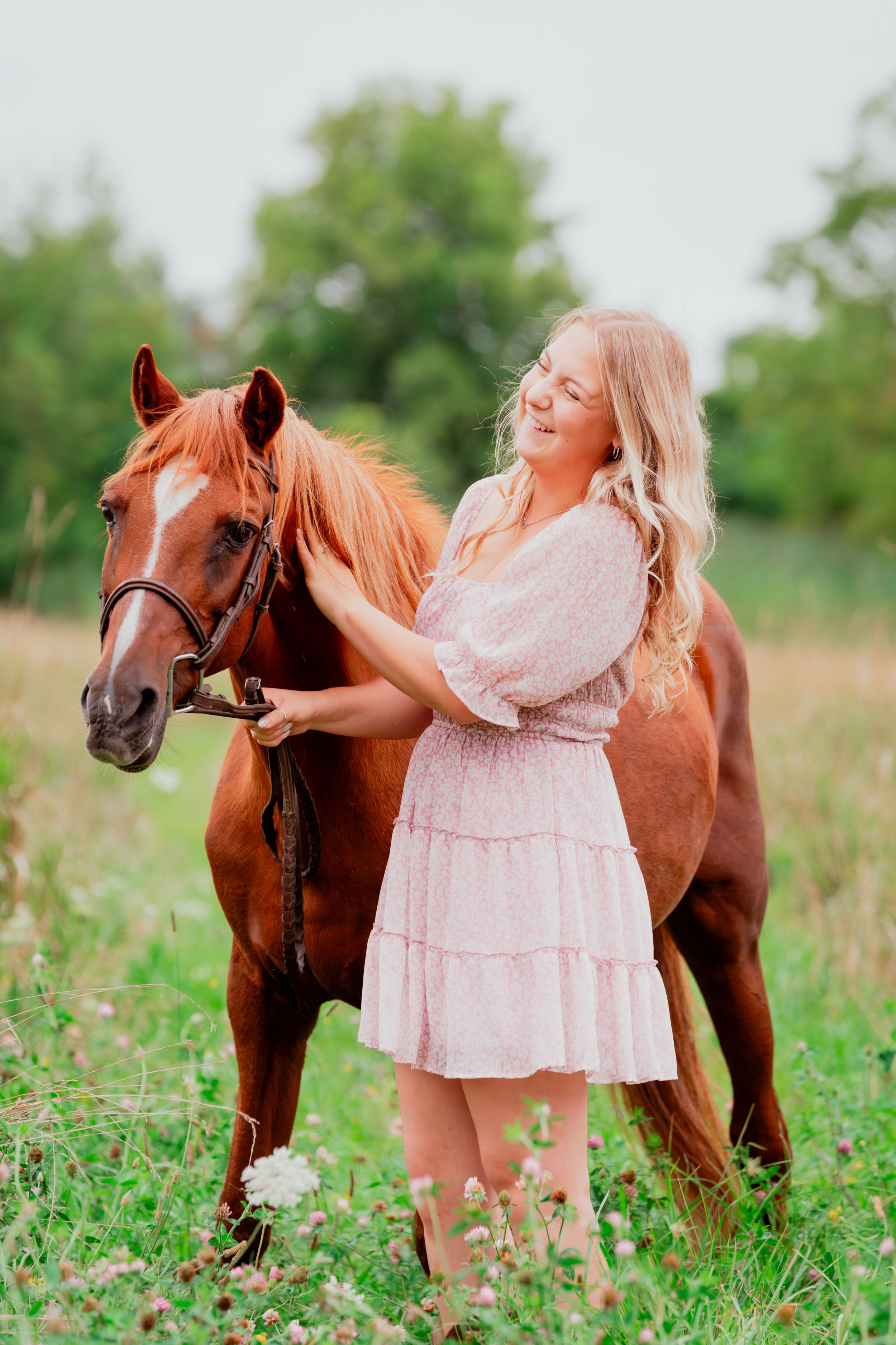 A person in a pink dress shares a happy moment with their chestnut horse in a flowery meadow.
