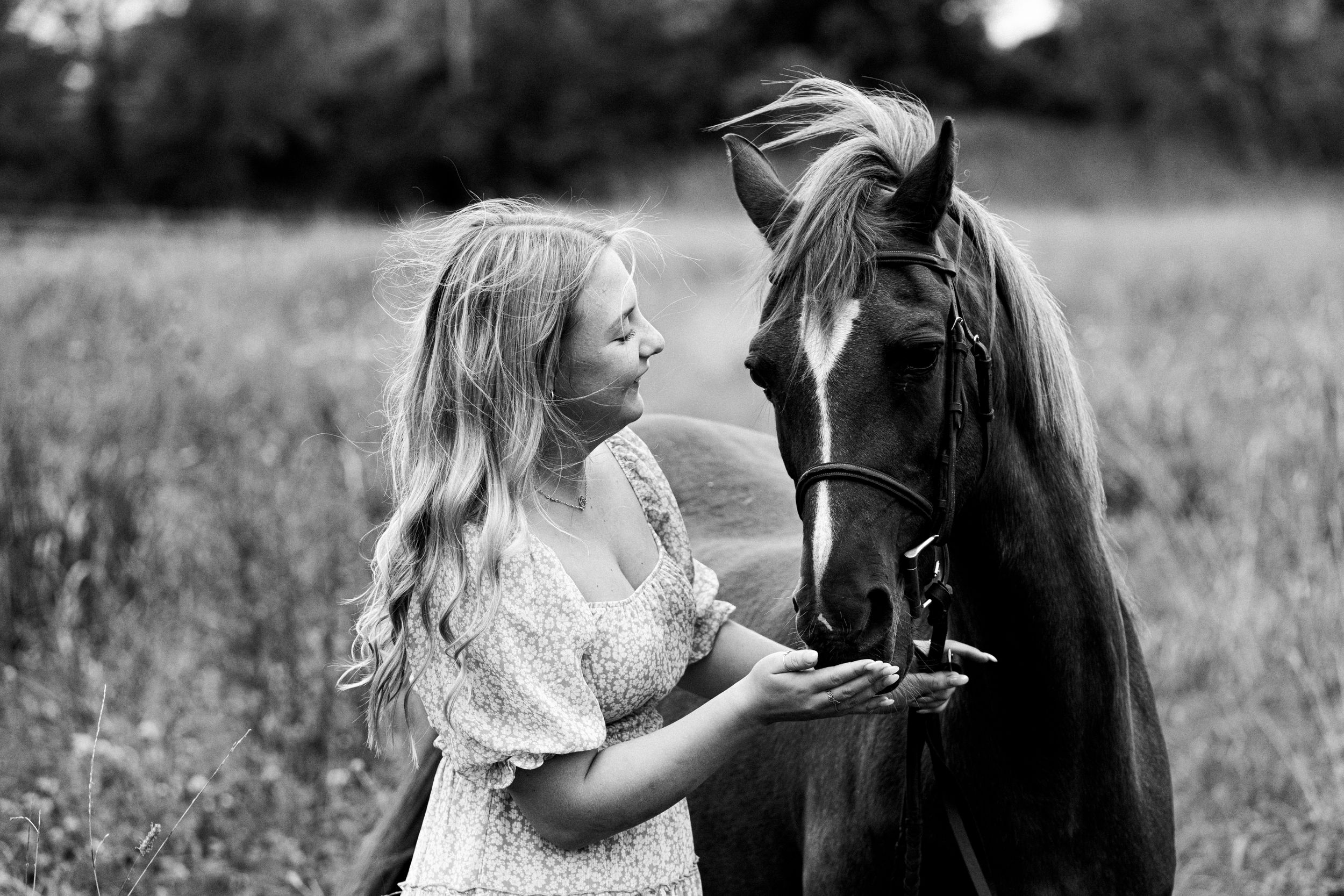 A black and white portrait showing a tender moment between a person and their horse in a field.