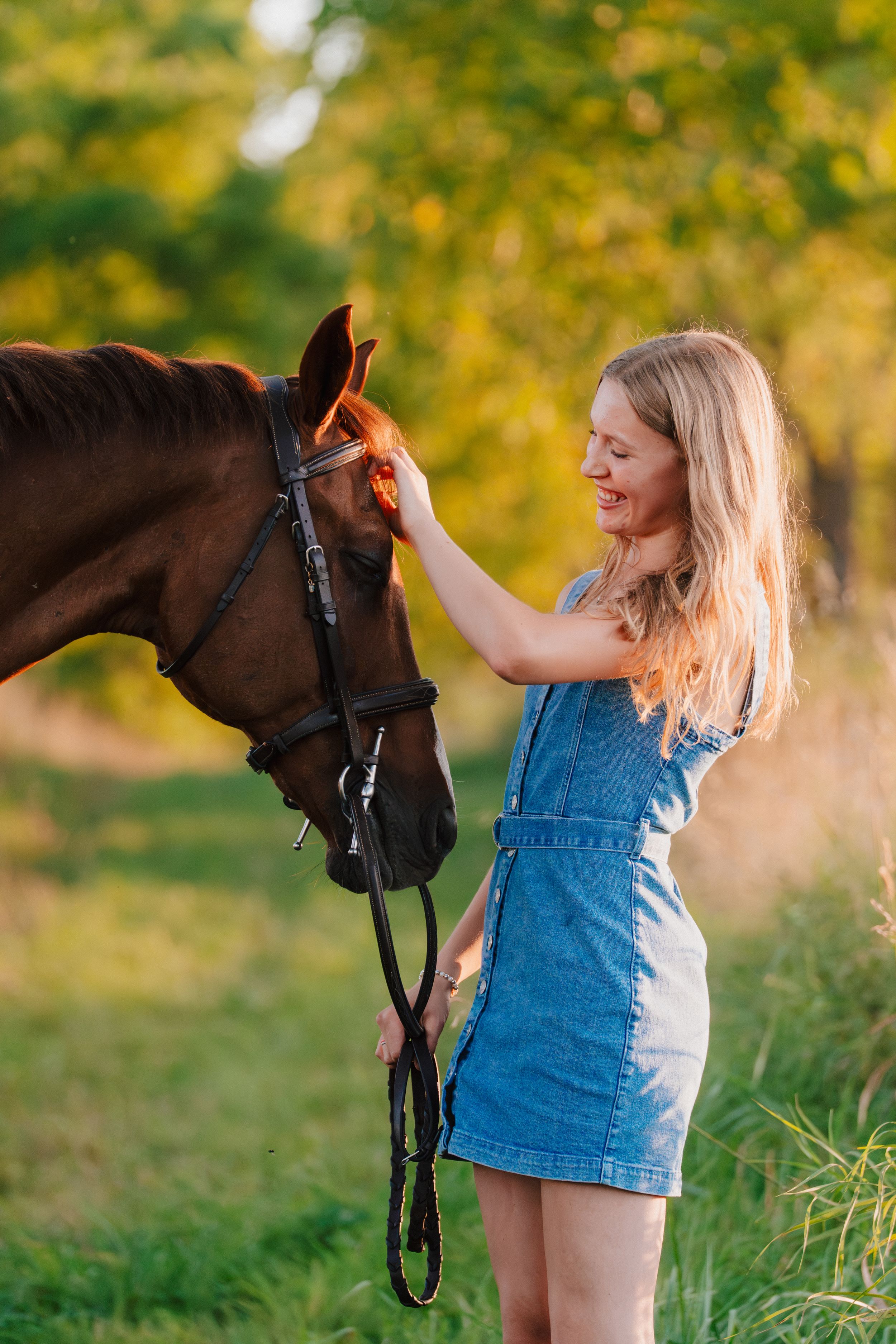 A person in denim overalls pets a brown horse in a sunny field at golden hour.