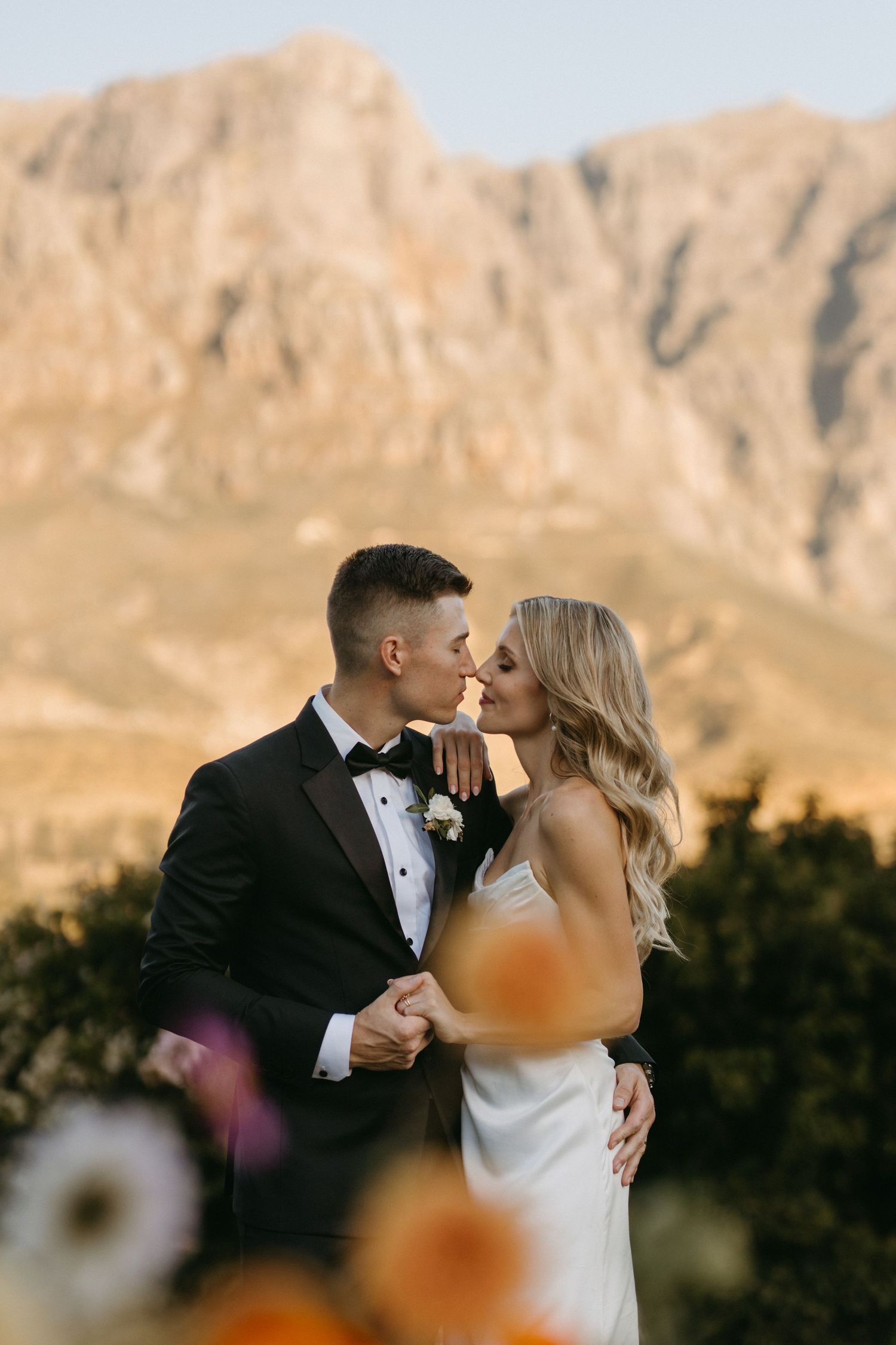 Bride and groom kiss against dramatic mountain backdrop at sunset.