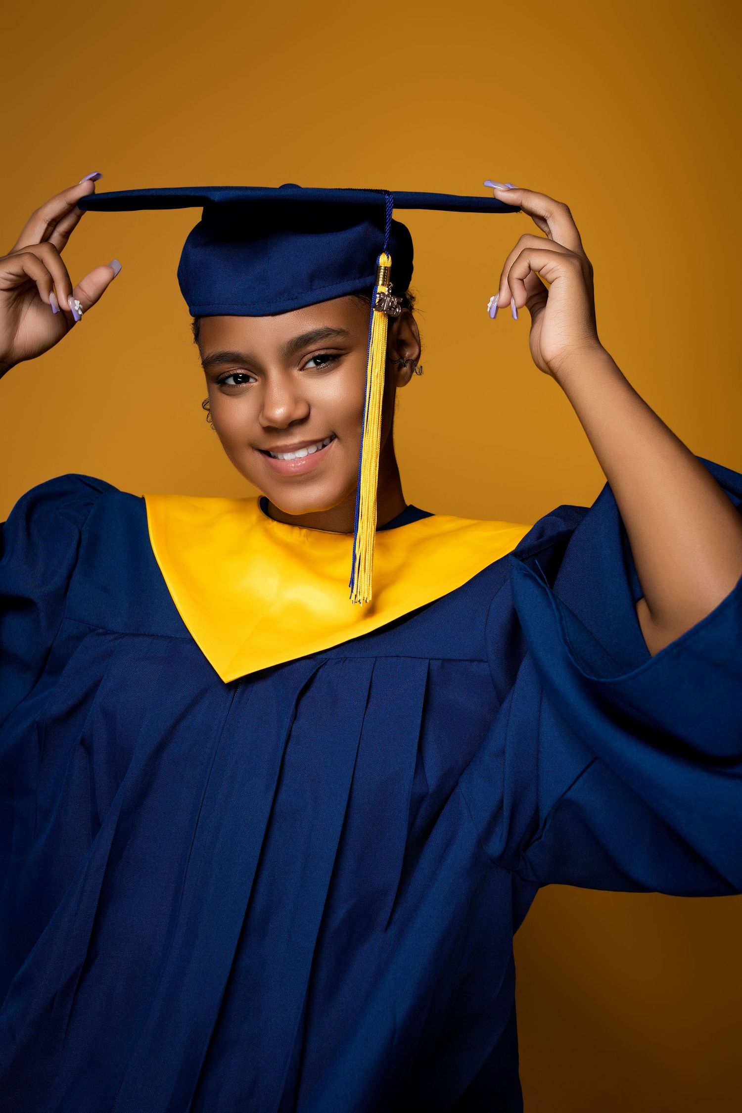 Graduate in blue and gold regalia poses confidently against orange backdrop during graduation photoshoot.