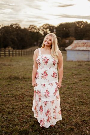 Smiling blonde woman in white floral maxi dress standing in a green field at golden hour near a rustic barn.