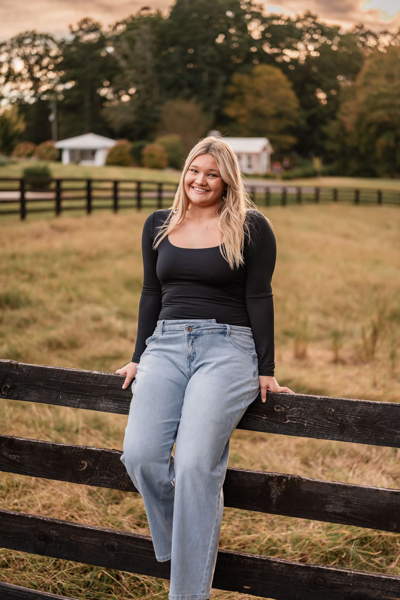 Blonde woman in black top and light jeans leaning on wooden fence at a farm during golden hour.