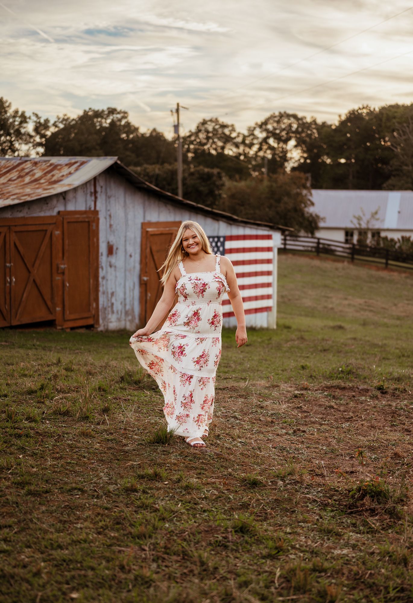 Blonde woman in floral maxi dress poses on farm with American flag barn backdrop at golden hour.