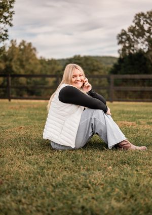 Smiling blonde woman sitting on grass in white vest and jeans, posing outdoors near a wooden fence.
