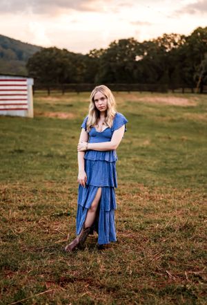 Country style senior portrait in tiered blue dress with American flag backdrop at sunset.
