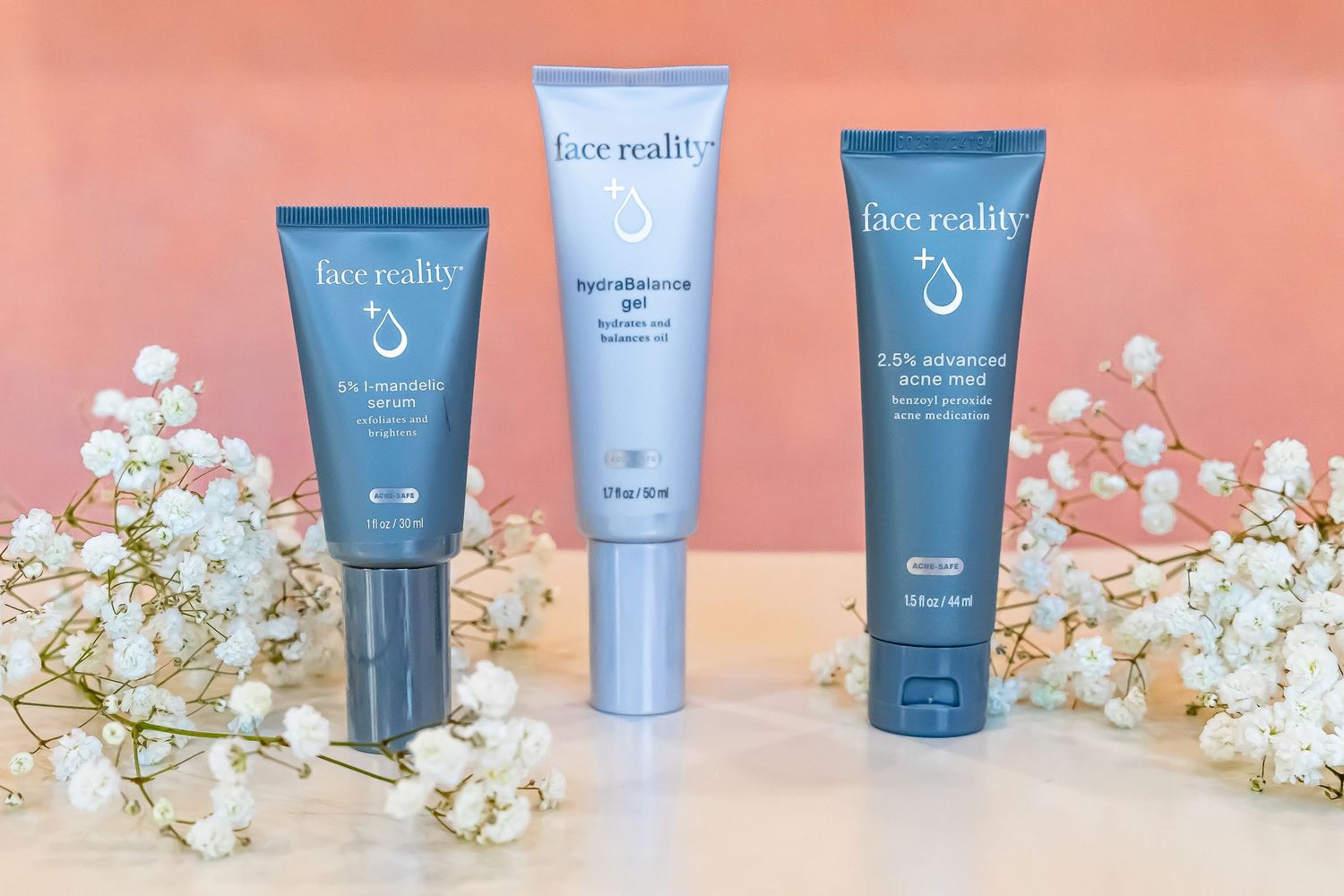 Three blue Face Reality skincare product tubes displayed on a coral background with delicate white baby's breath flowers.