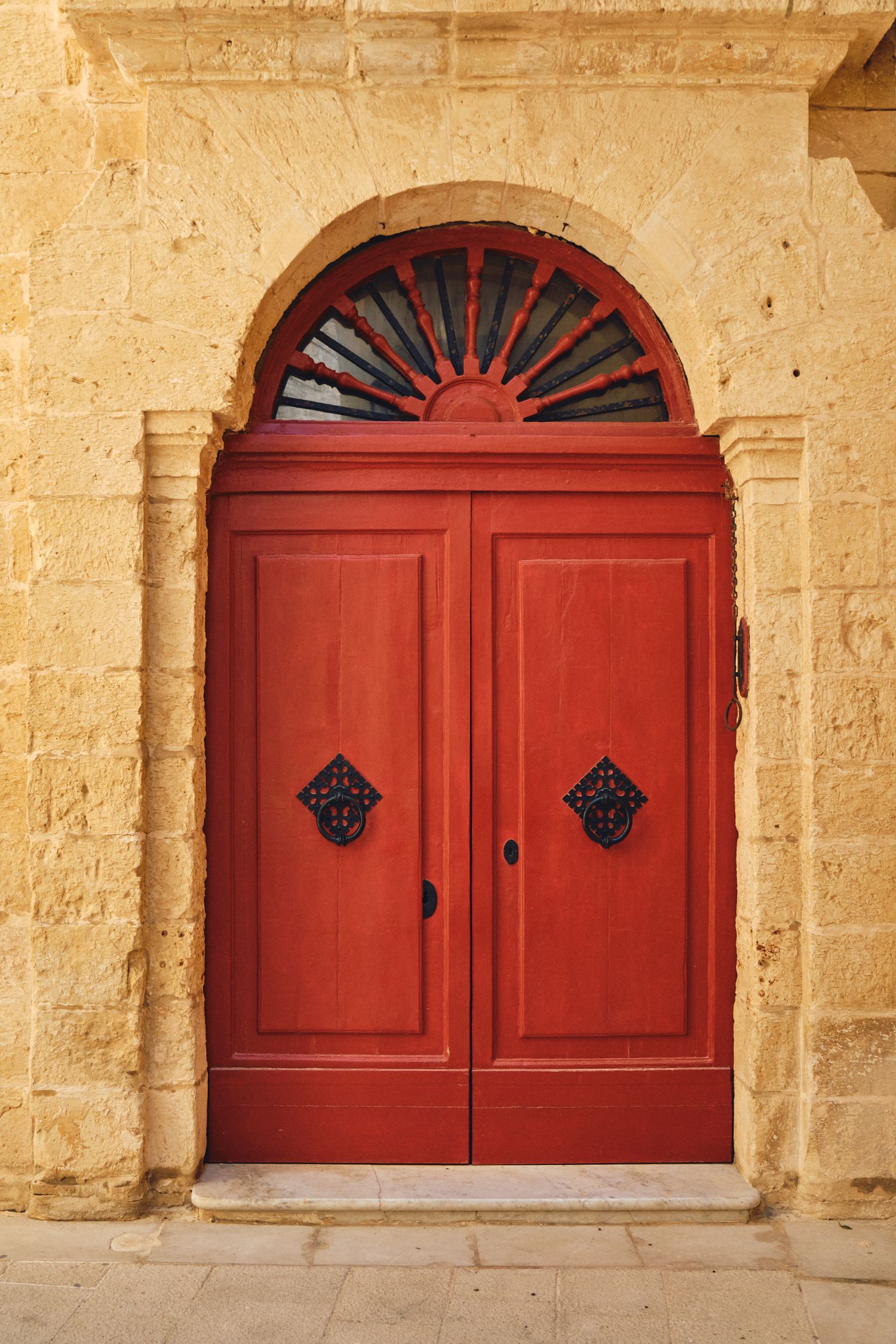 Red double doors with fanlight window and ornate hardware set in limestone archway.