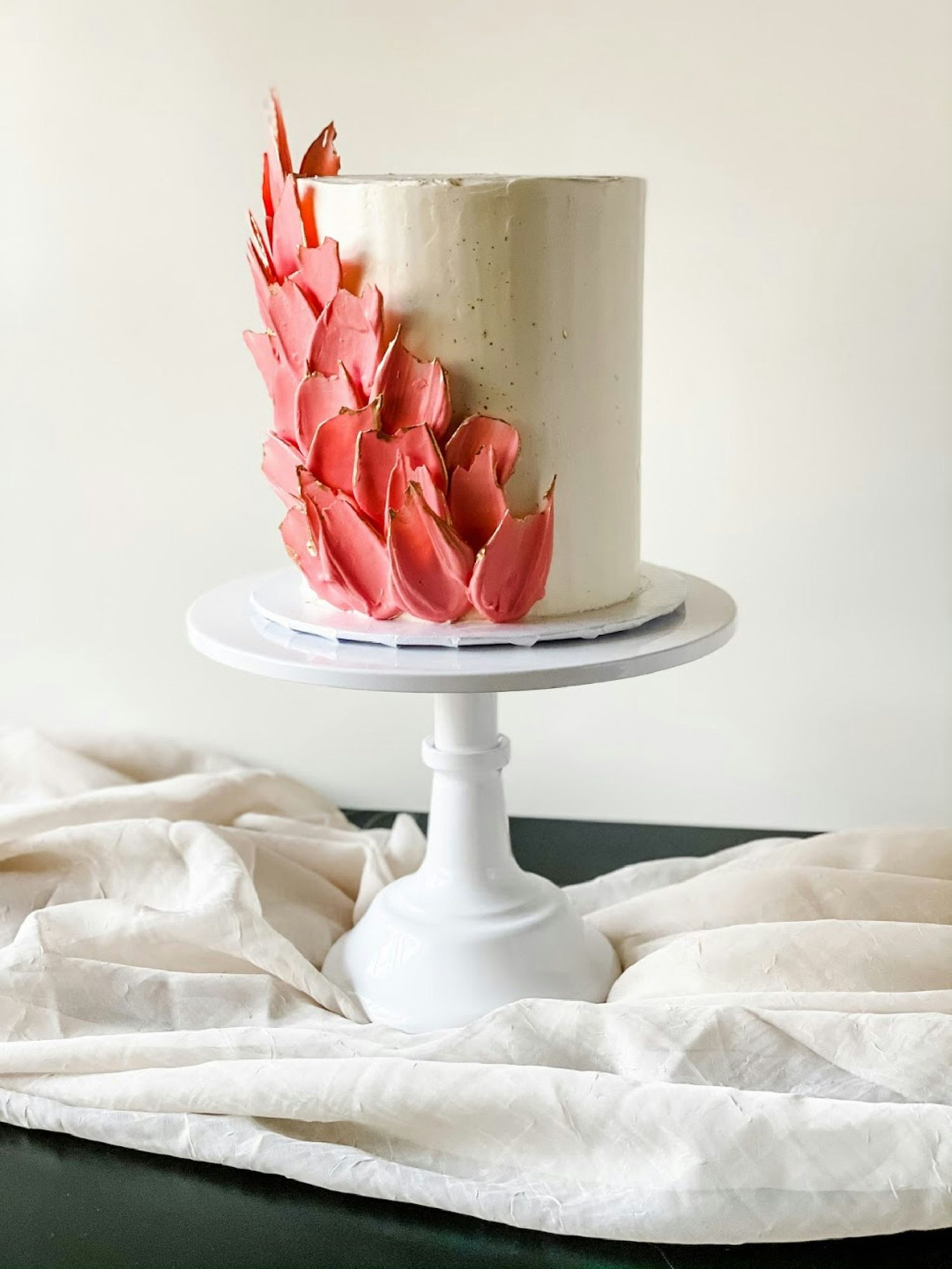 Elegant white wedding cake decorated with cascading coral-colored sugar petals on a classic white pedestal stand.