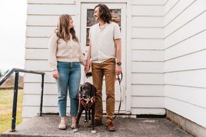 A couple stands with their black dog outside a white building on a cloudy day.