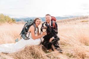 A bride and groom in backpacking gear kneel beside their Bernese Mountain Dog in a field.