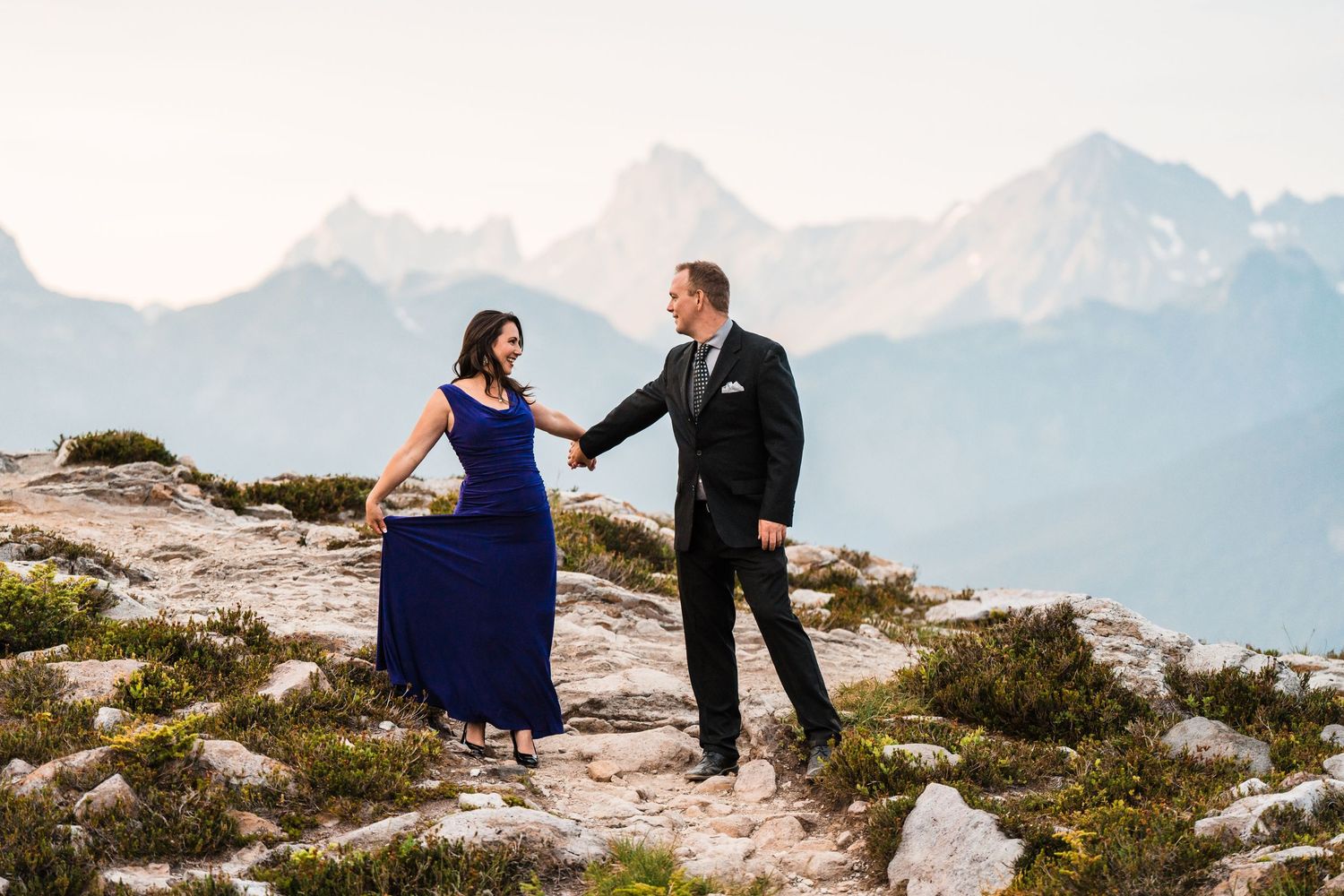 Couple in formal wear holding hands on a rocky mountain ridge with snowy peaks behind.